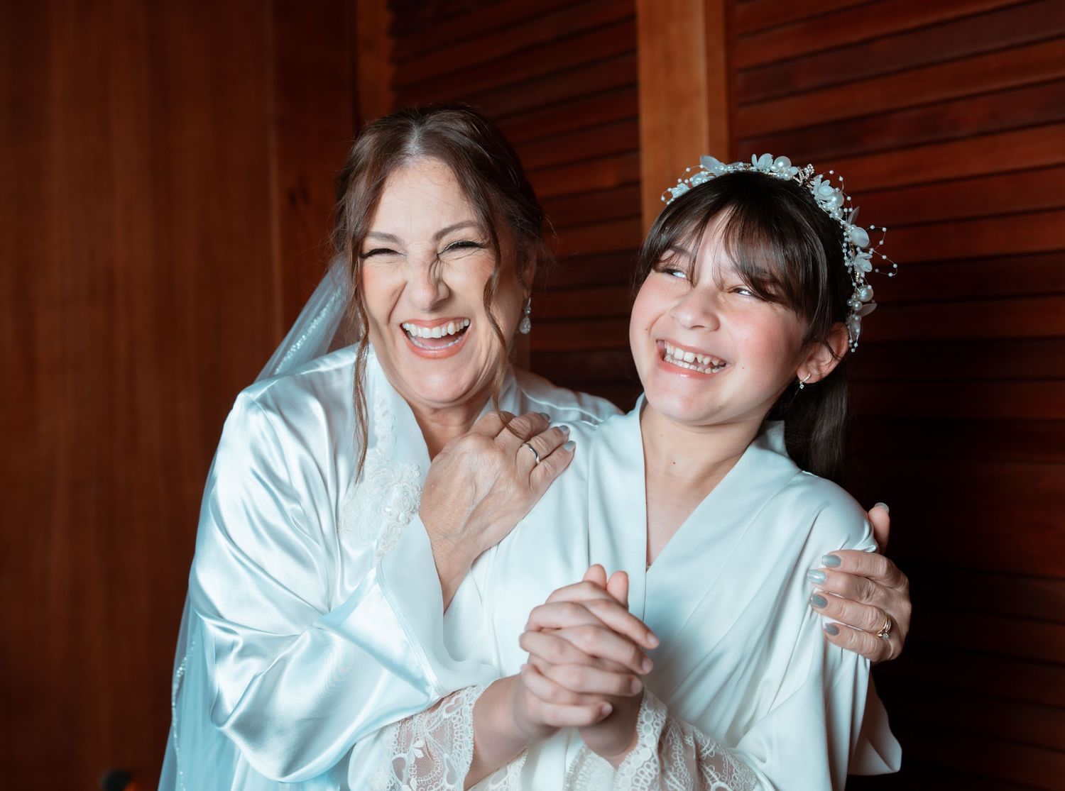 Two people in white satin robes share a joyful moment while getting ready for a special occasion.