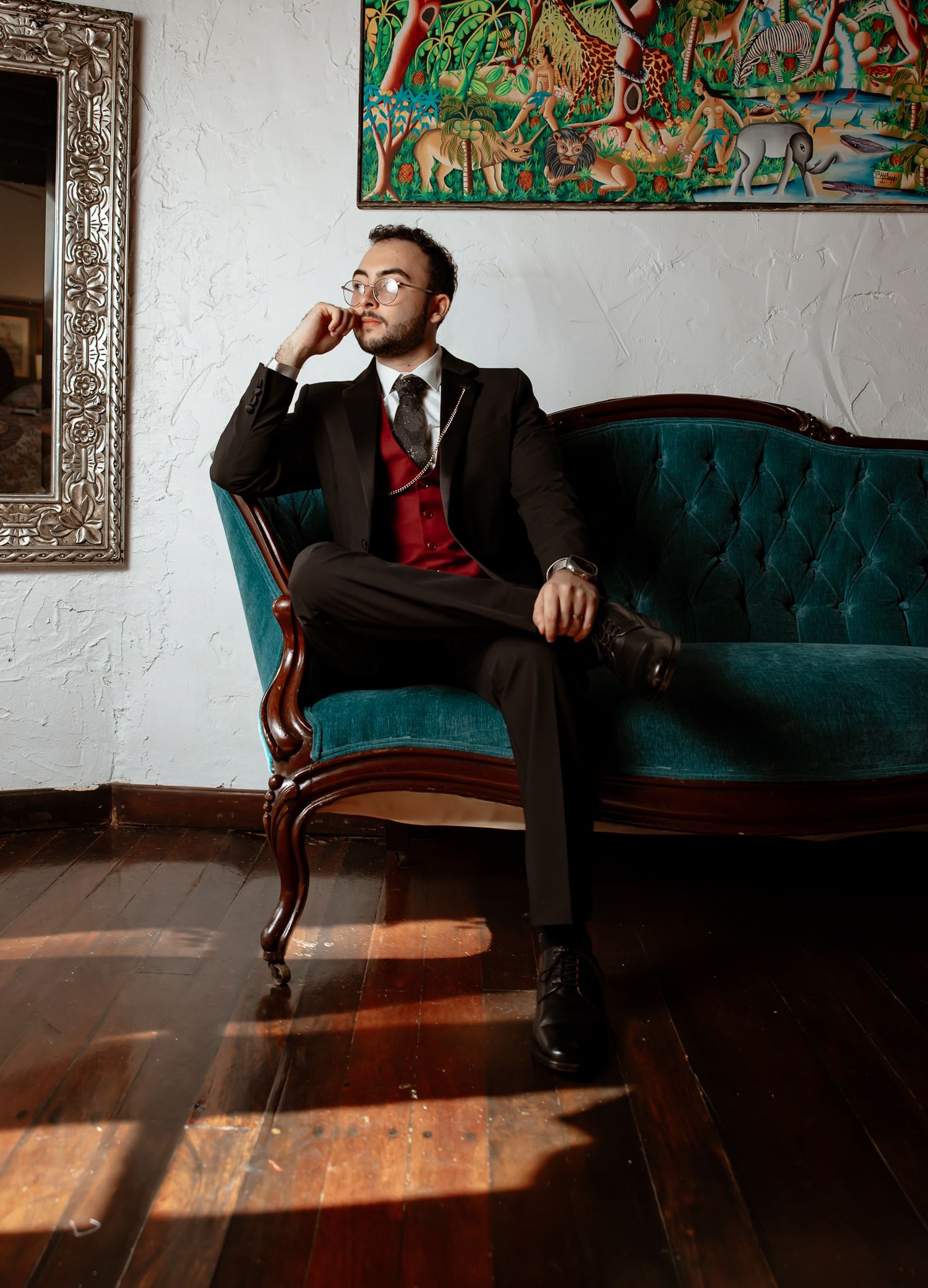 Elegant groom in black suit with red vest sits on tufted teal velvet sofa in sunlit room.