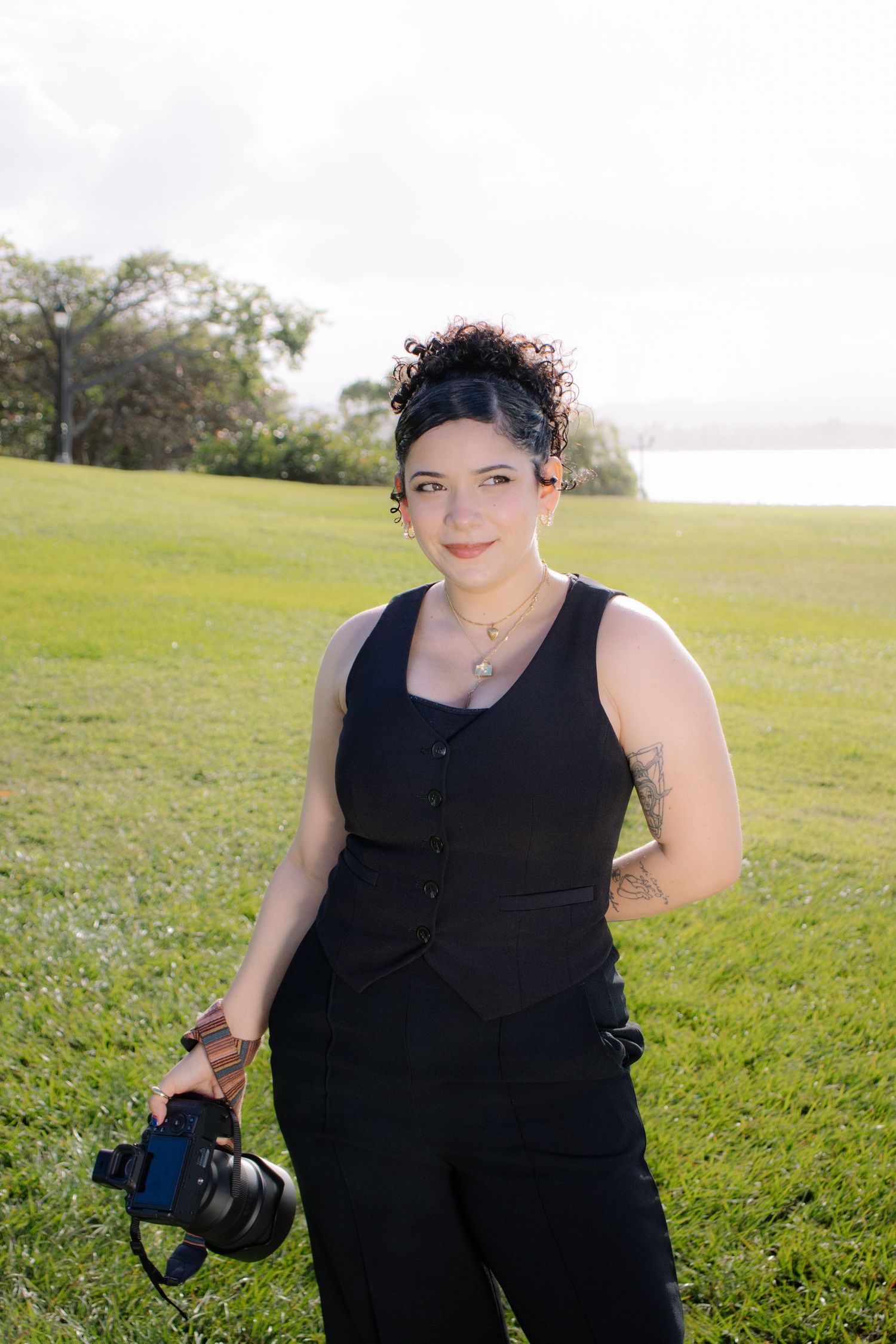 Professional photographer in black outfit poses with camera equipment on grassy waterfront.