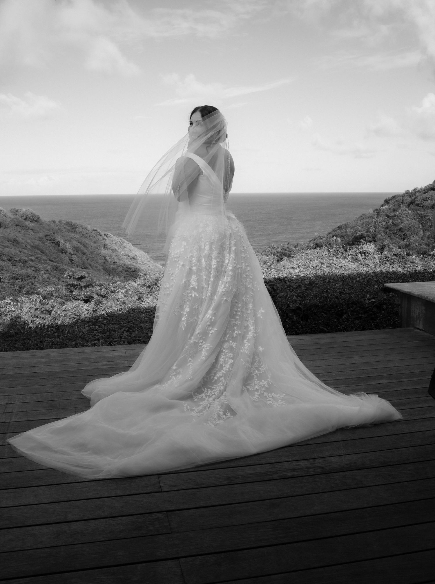 A bride in a flowing white lace wedding gown stands on a wooden deck overlooking a dramatic coastal landscape.
