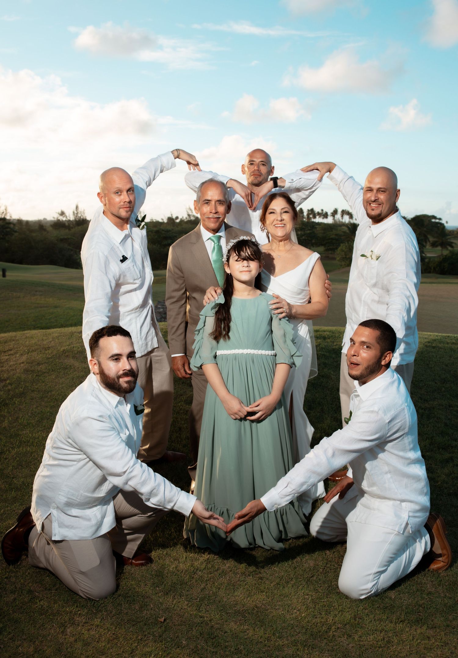 Wedding party members in mint green dresses and white suits pose playfully on grassy field during sunset.