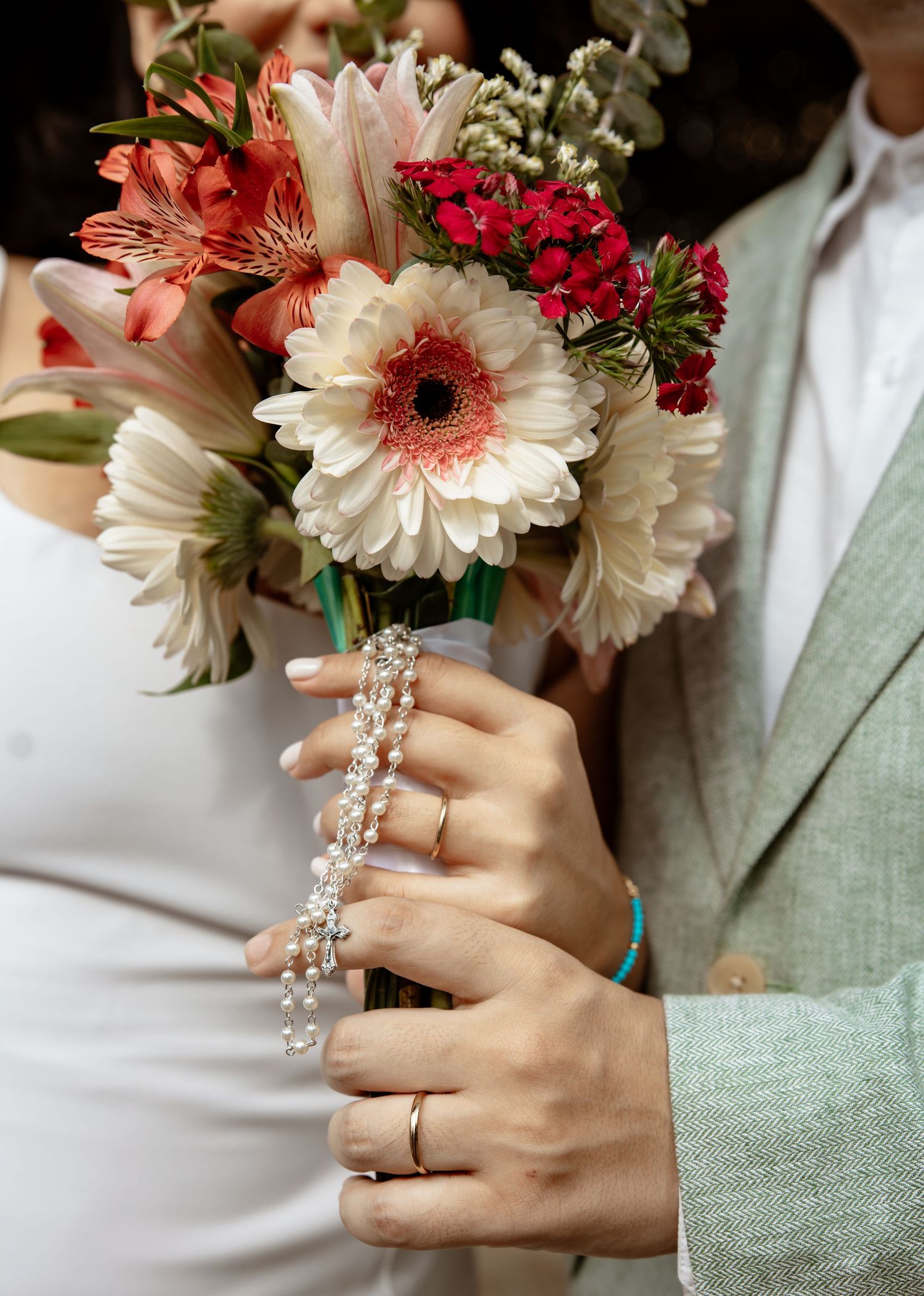 A bridal bouquet with pink gerbera daisies and crystal details is held by hands wearing rings.
