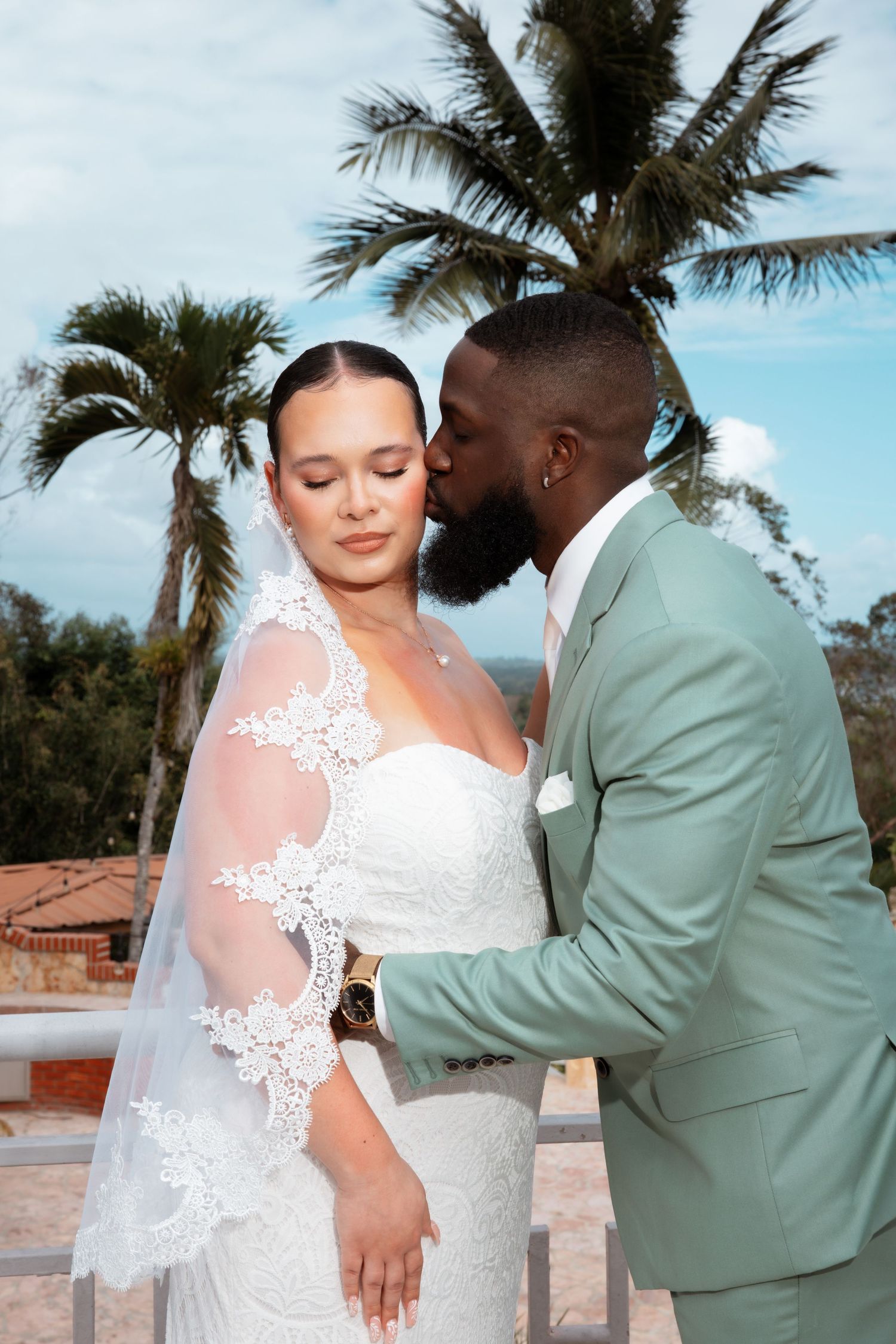 A romantic wedding couple embraces under palm trees, with the bride in lace and the groom in a mint green suit.