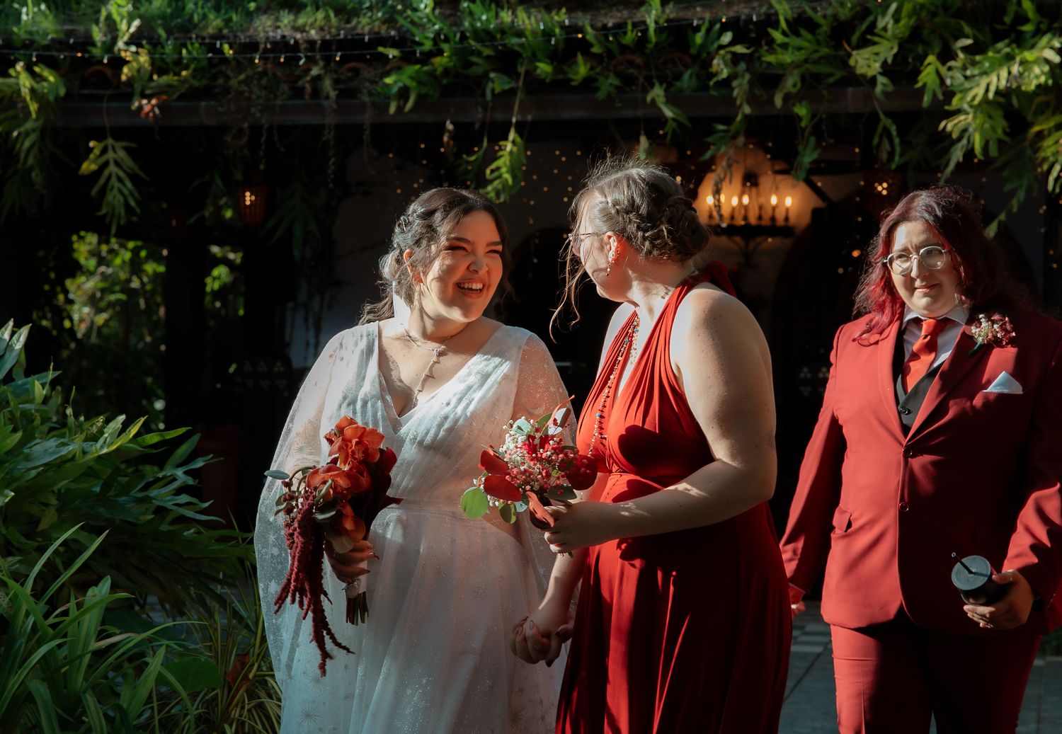 Bride in white gown laughing with bridesmaid in red dress and guest at elegant indoor wedding ceremony.