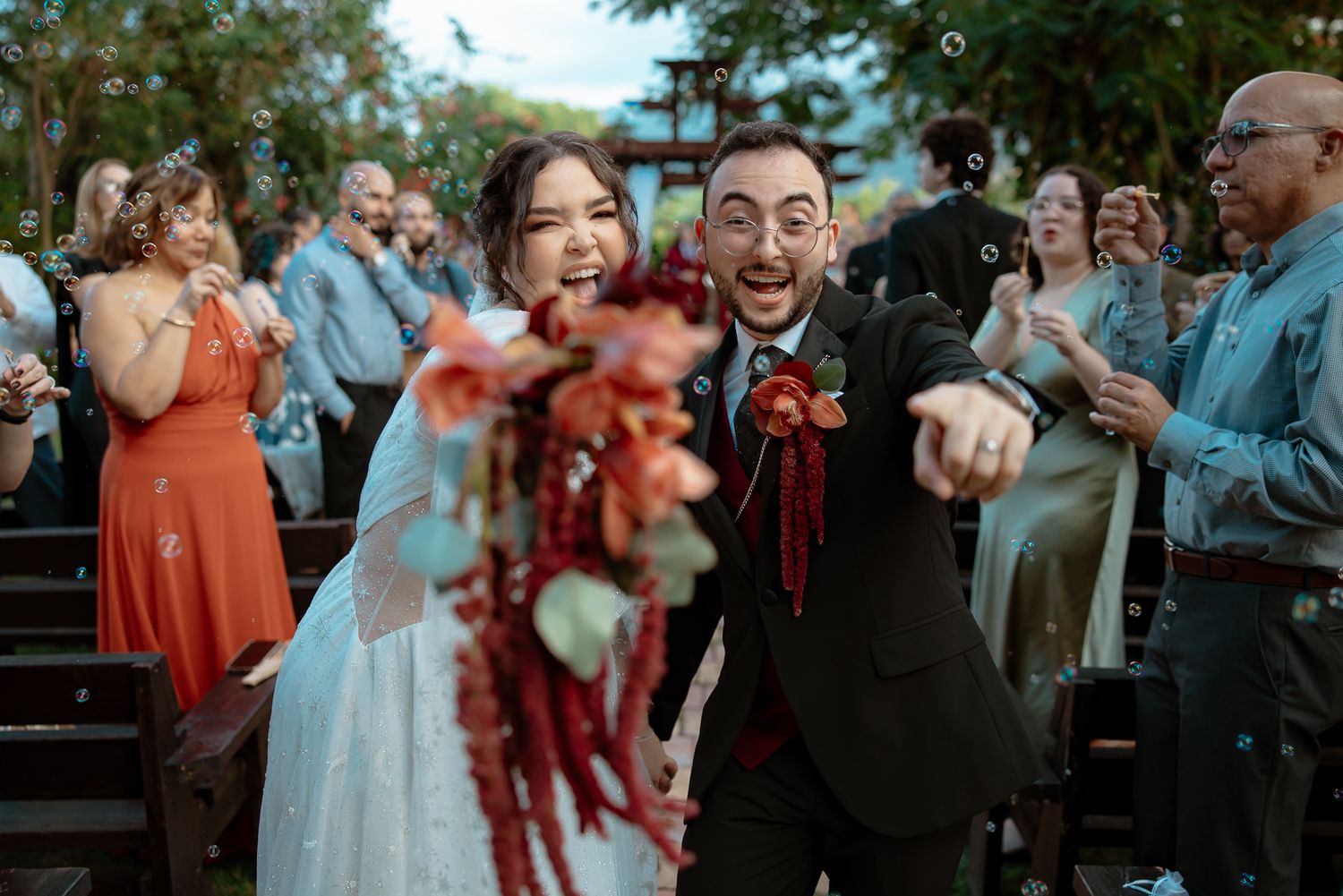 Joyful newlyweds walk down the aisle surrounded by bubbles, bride holding red bouquet, guests celebrating outdoors.