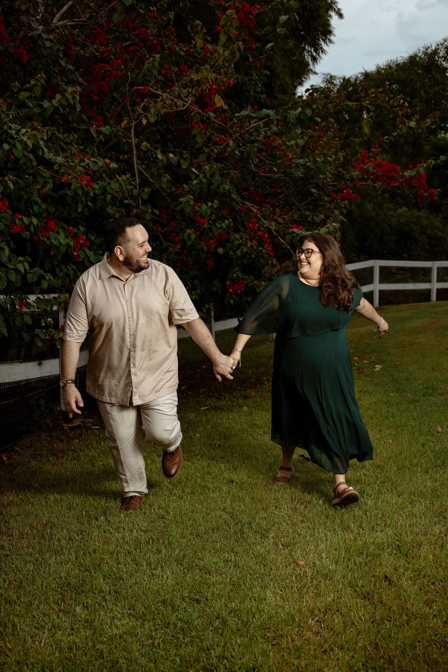 Couple playfully dancing together on a grassy lawn with red flowering trees in the background.