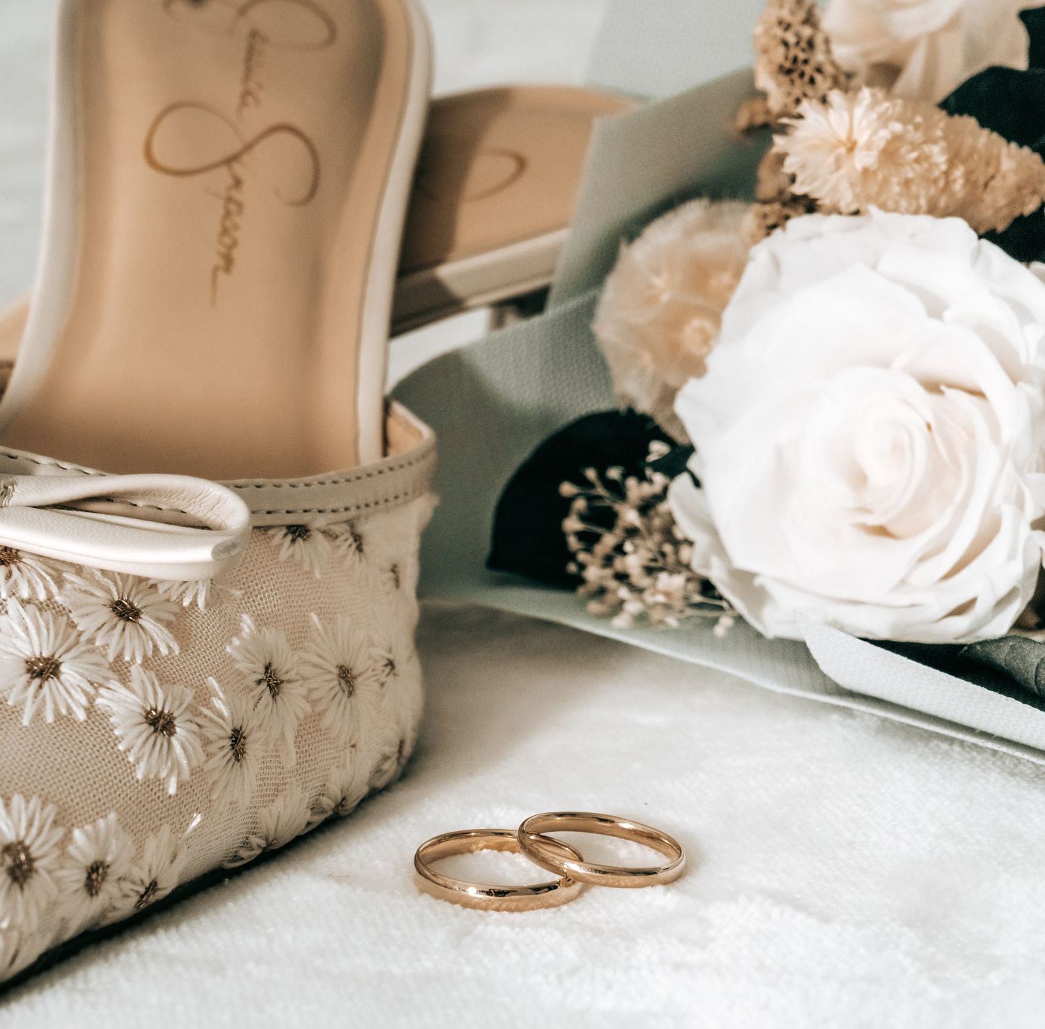Elegant wedding shoes and rings arranged with delicate roses on white surface.