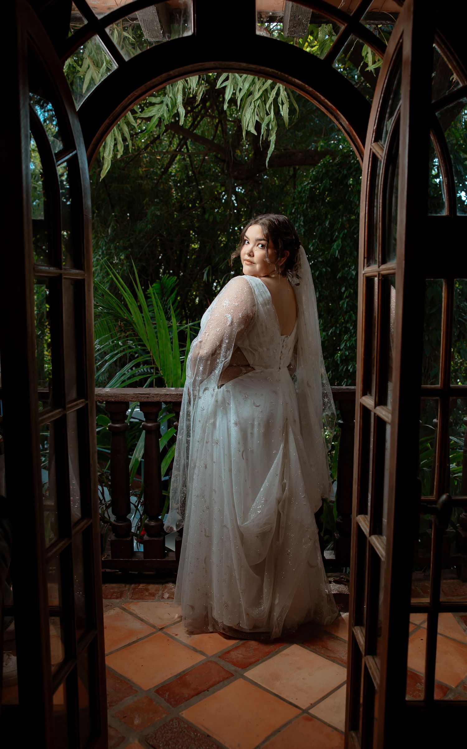 Bride in white lace gown and veil posing in garden archway at outdoor wedding venue.