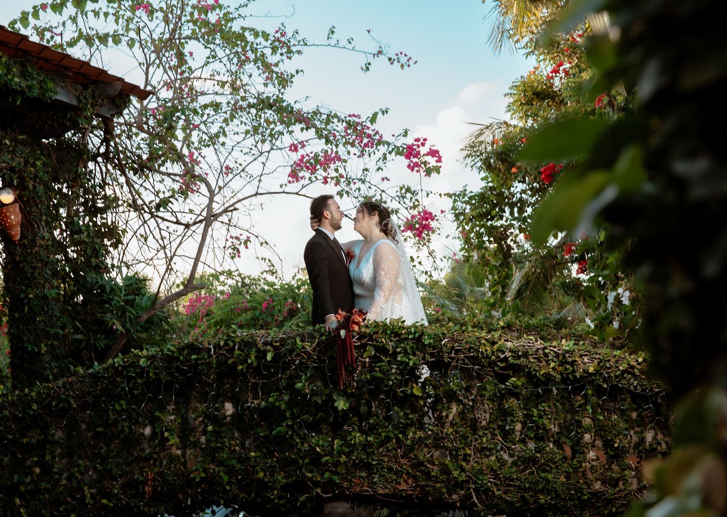 Wedding couple poses in a lush garden setting with flowering bushes and natural greenery.