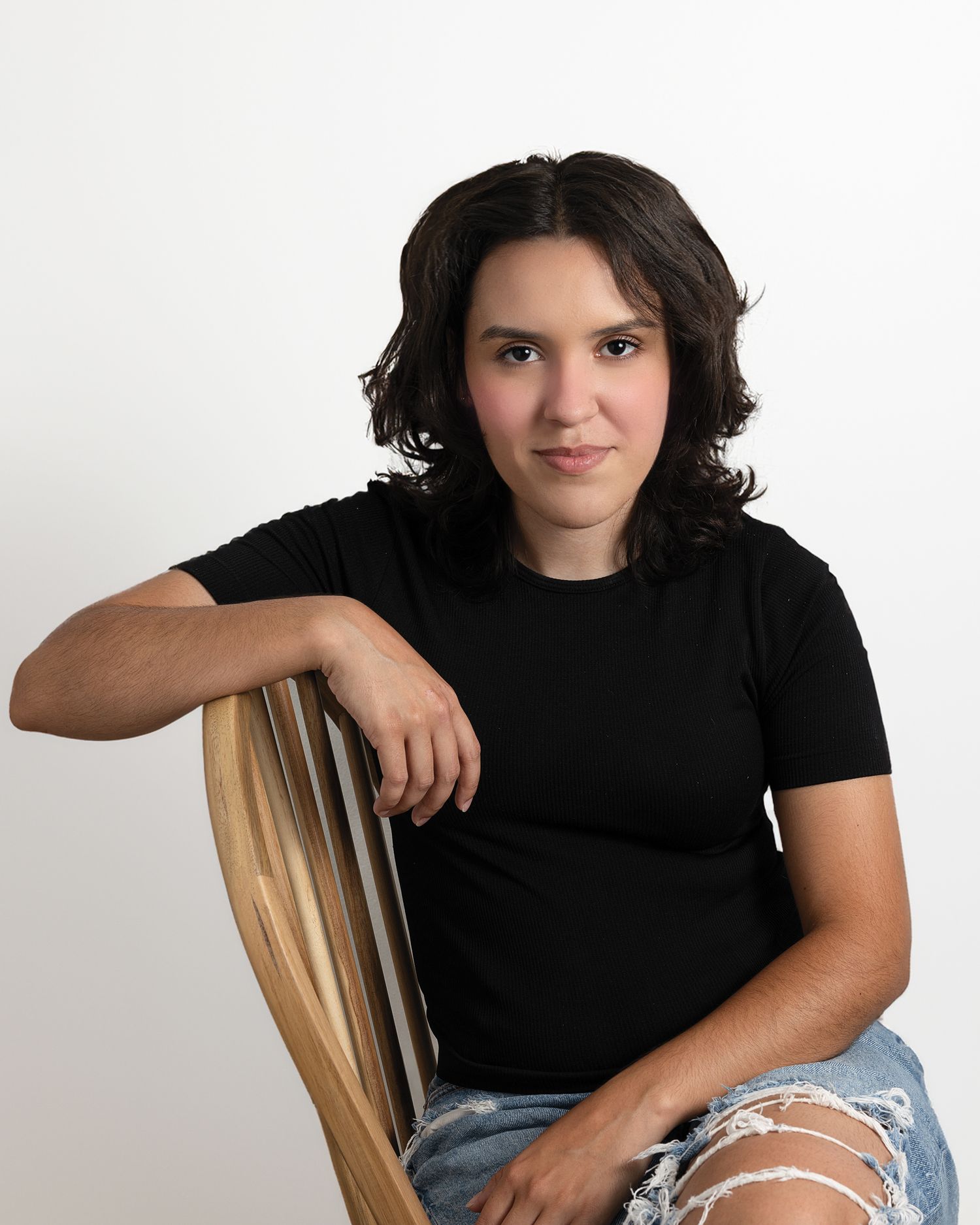 Person in black t-shirt and ripped jeans sitting casually on wooden chair for acting headshot.