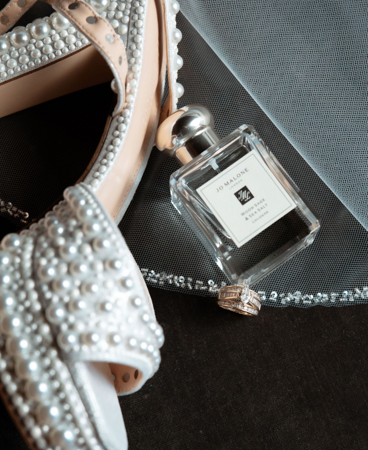 Close-up details of elegant wedding accessories showing perfume bottles and pearl-adorned bridal shoes on dark background.