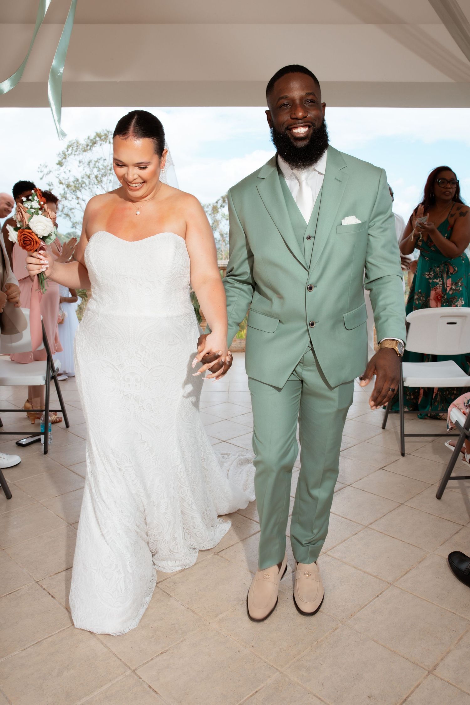 A bride in a white lace gown and groom in a mint green suit dance together on a tiled floor at their wedding reception.