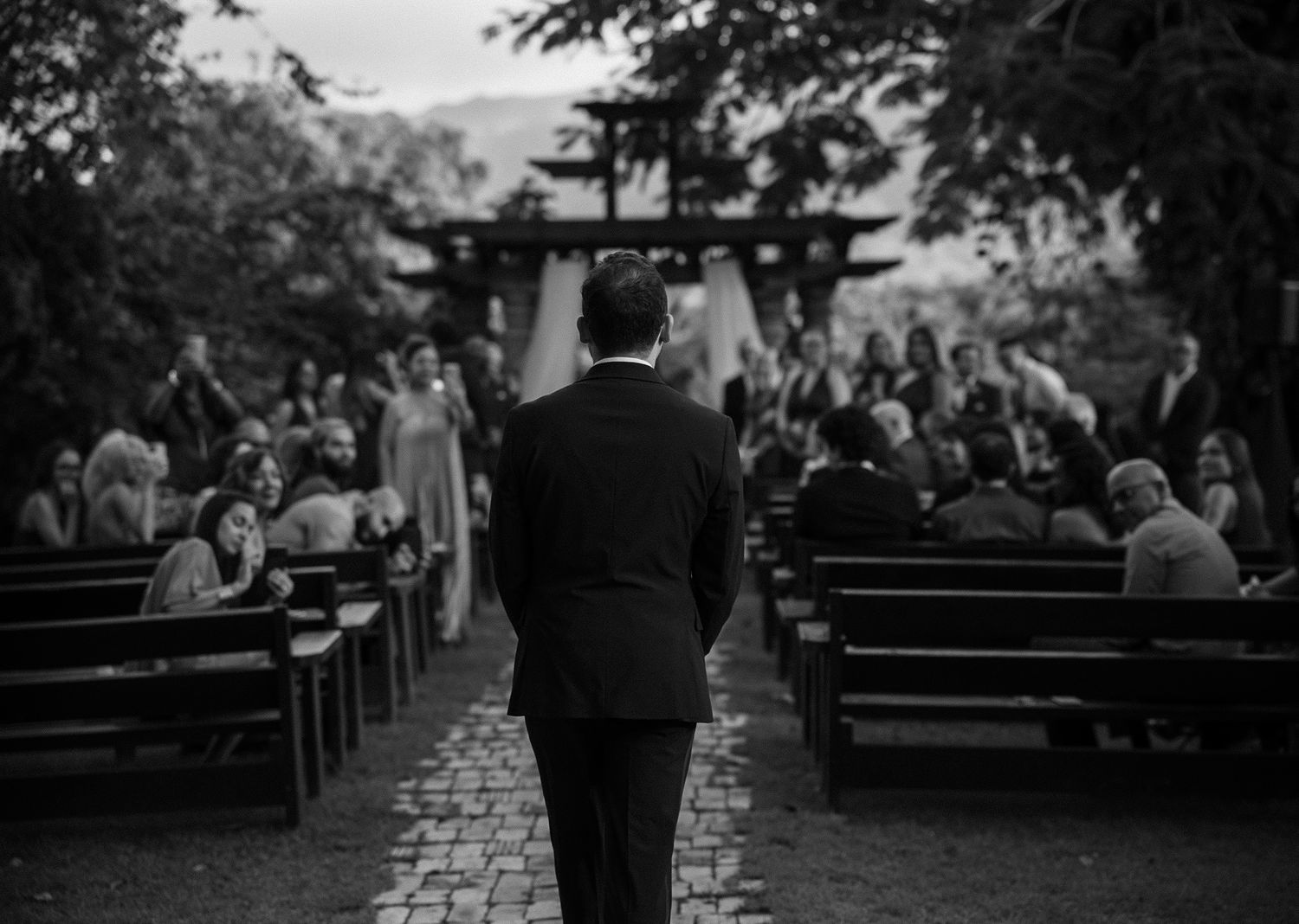 Groom walks down outdoor wedding aisle lined with wooden pews and guests in black and white photo.