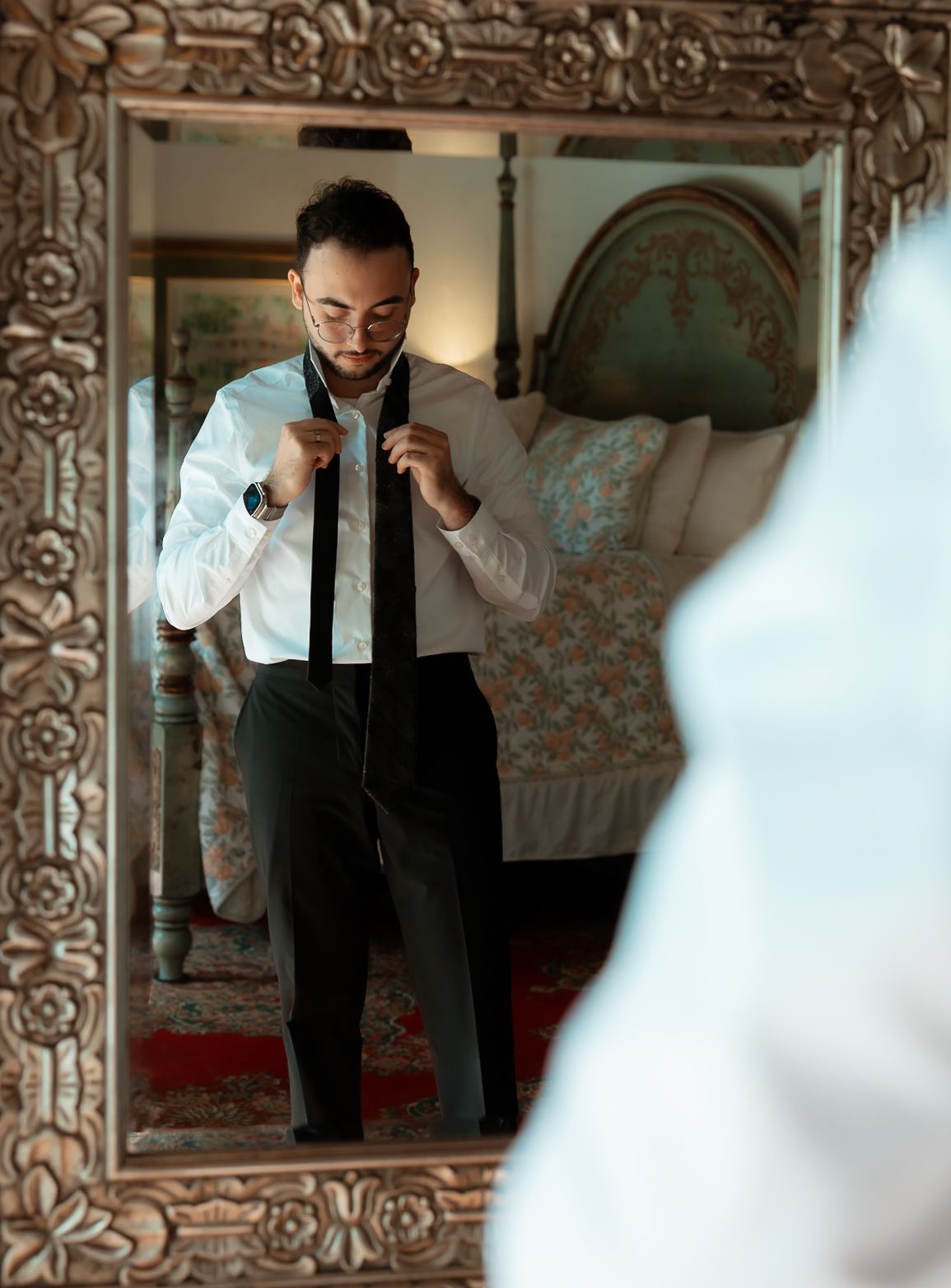 Groom adjusting black tie in ornate mirror, wearing white shirt and suspenders in elegant bedroom.