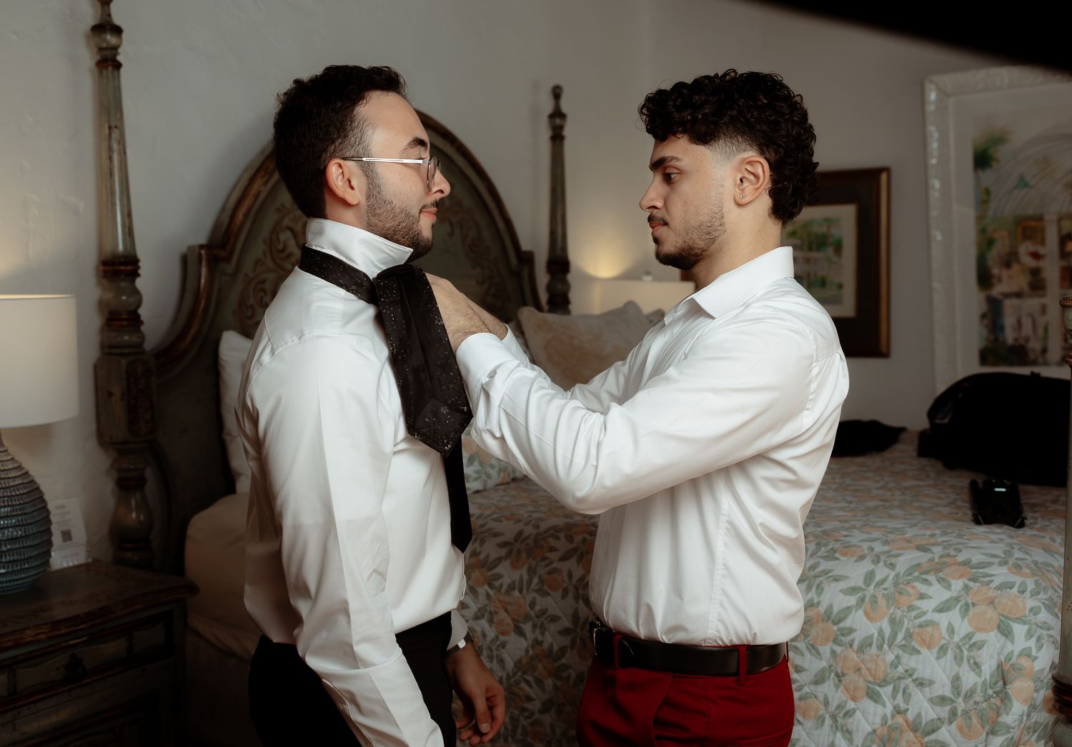 Groomsman helps adjust the tie of a man wearing glasses in a bedroom during wedding preparations.