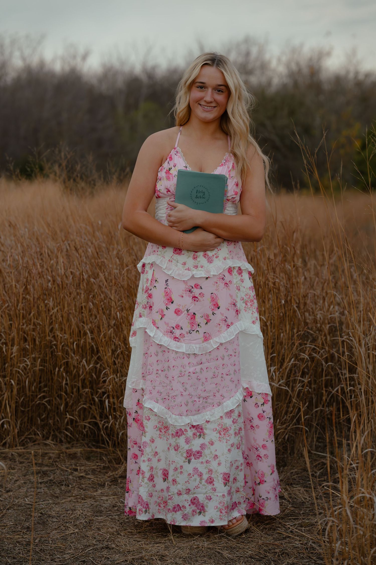 Young woman in pink floral maxi dress holding teal book, standing in golden field at dusk.