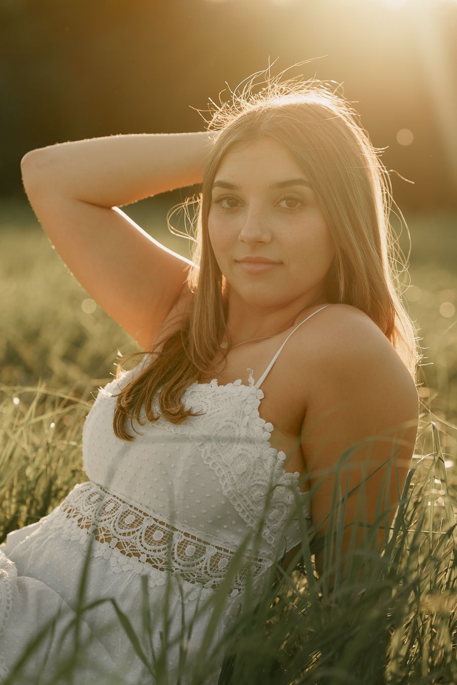 Young woman in white lace sundress sitting in green field, golden hour backlit portrait with hand in hair.