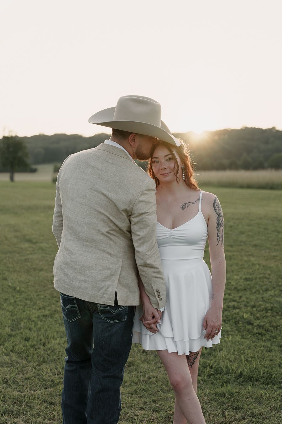 Couple holding hands at sunset in a green field, man in cowboy hat and blazer, woman in white mini dress with tattoos.