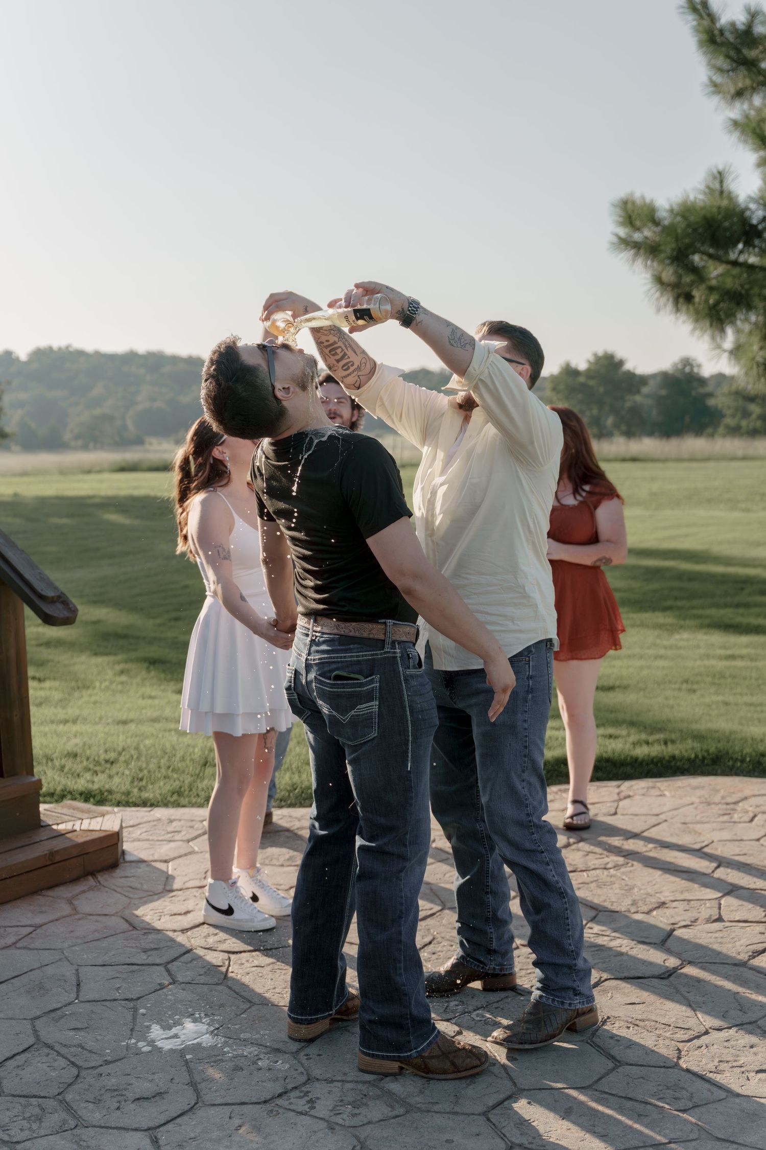 Group of friends toasting drinks outdoors on a patio at sunset, with green fields in the background.