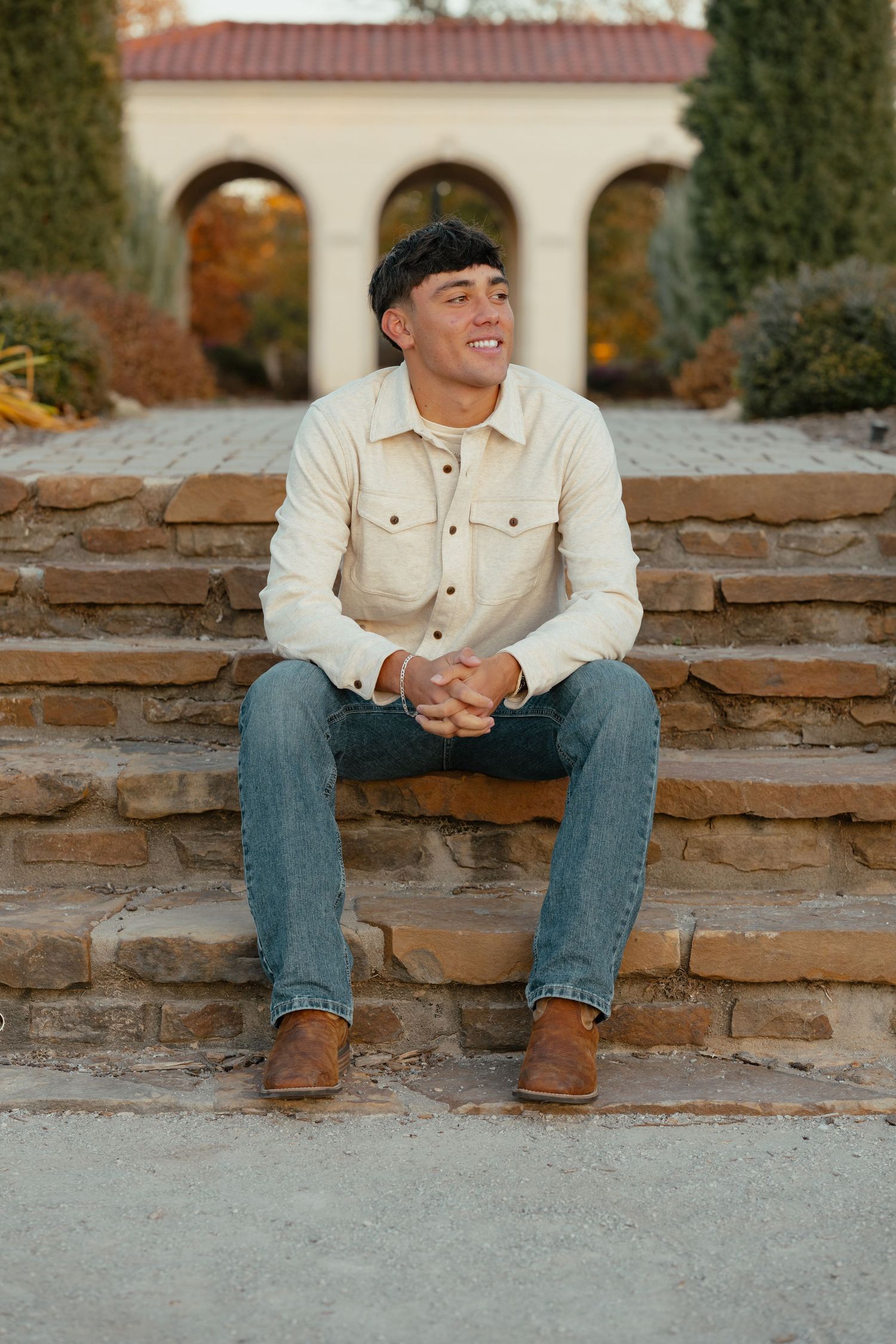A person in a beige jacket and jeans sits casually on stone steps during an outdoor photo session.