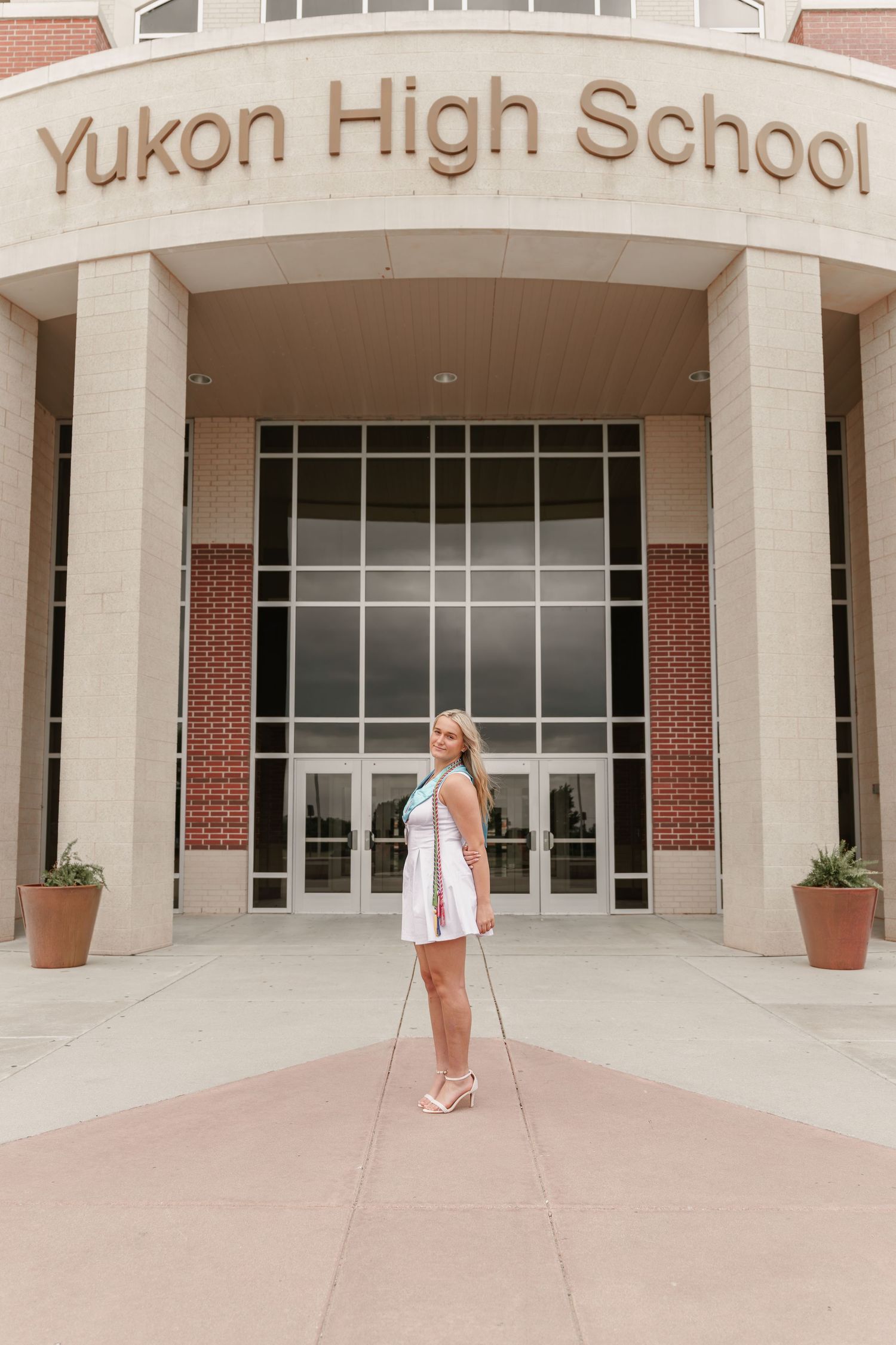 A young woman in a white dress poses in front of Yukon High School entrance for senior photos.
