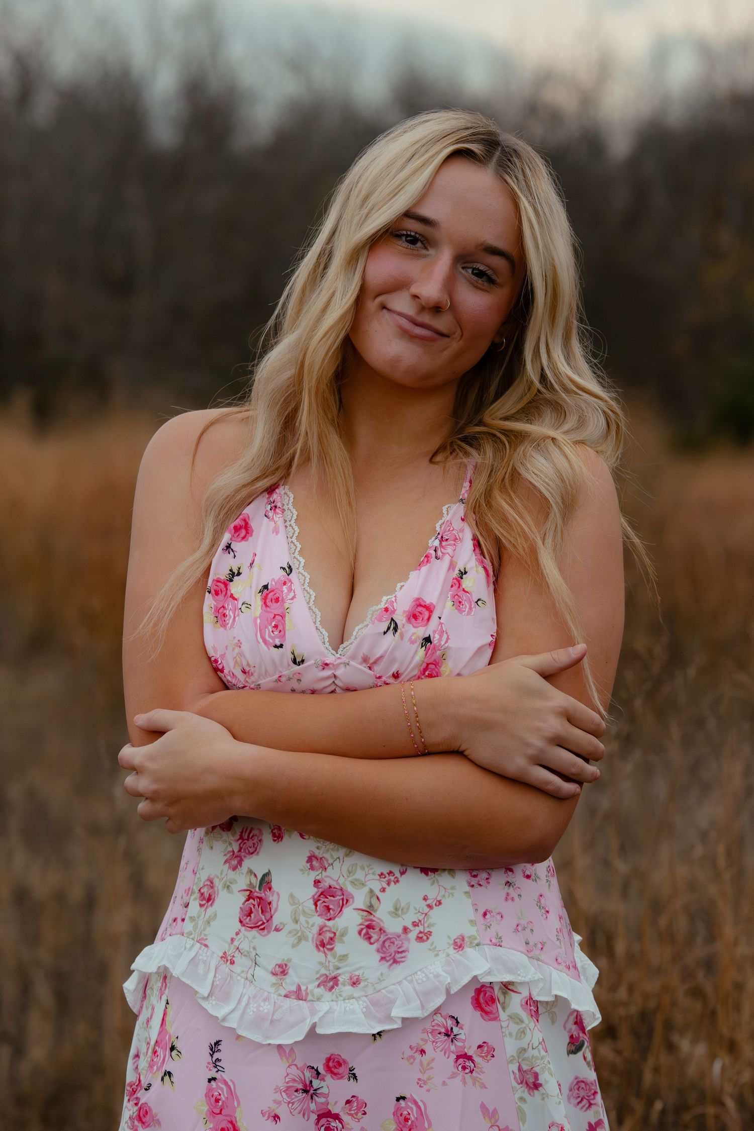 Young blonde woman in pink floral dress posing outdoors in a field with arms crossed, smiling softly.