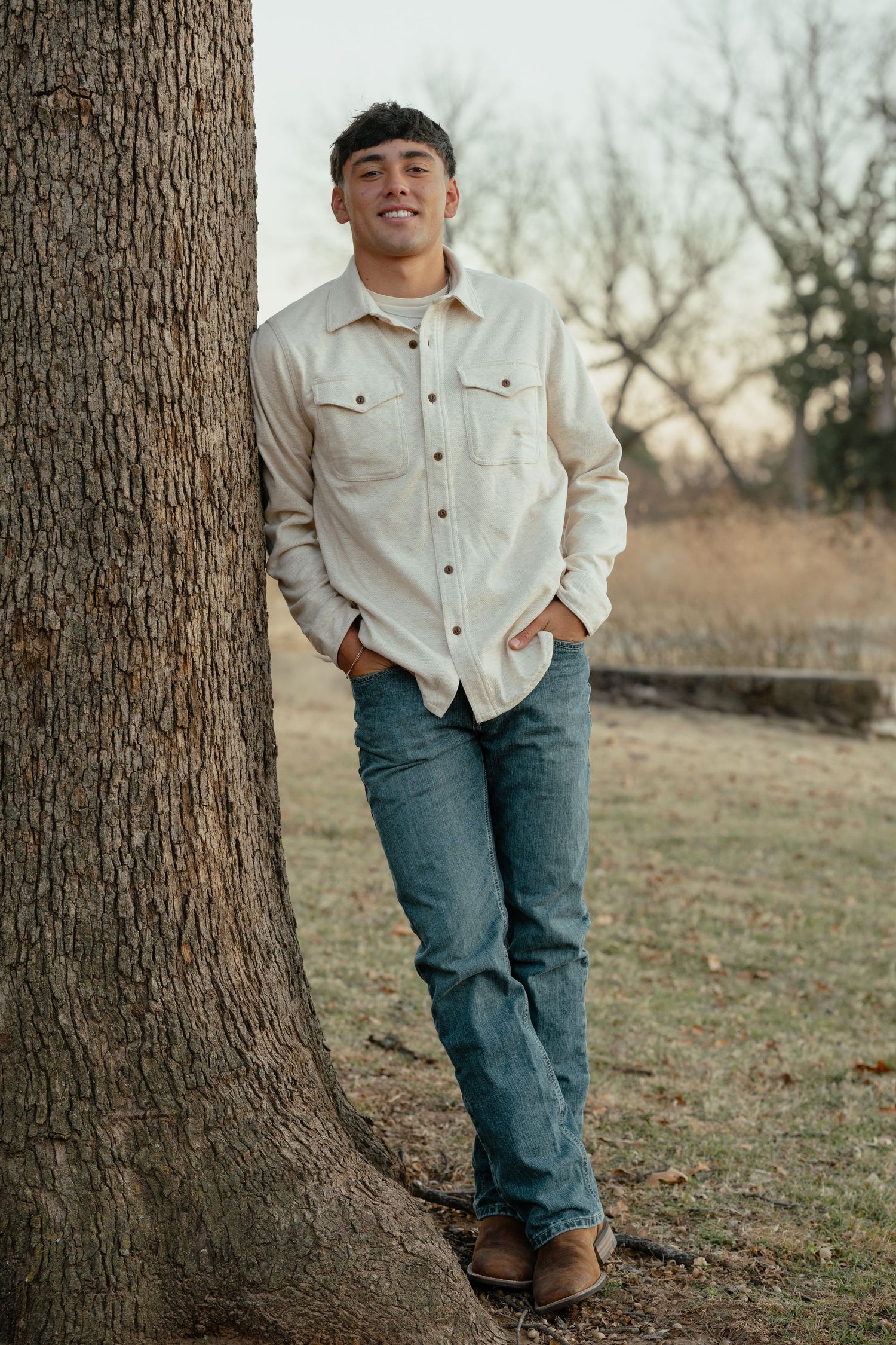 Person in casual fall attire poses next to large tree trunk in an outdoor setting.