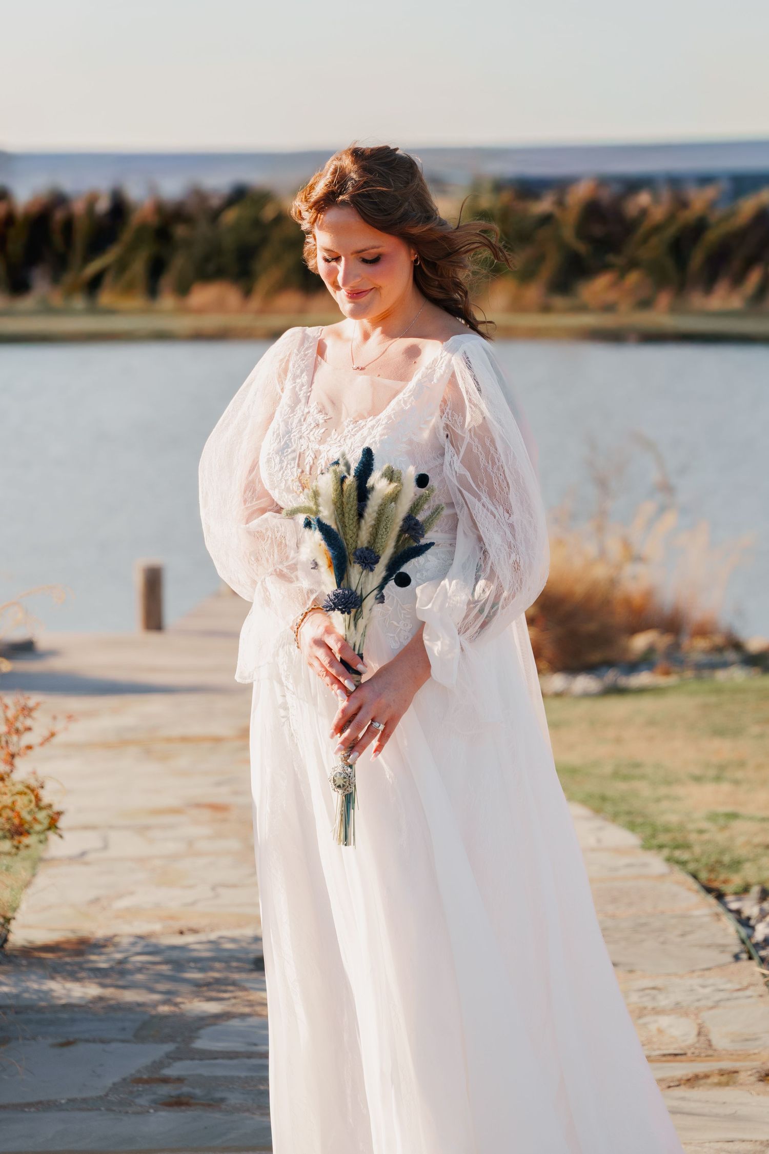Bride in white lace gown with puff sleeves holds dried flower bouquet outdoors near a lake at golden hour.
