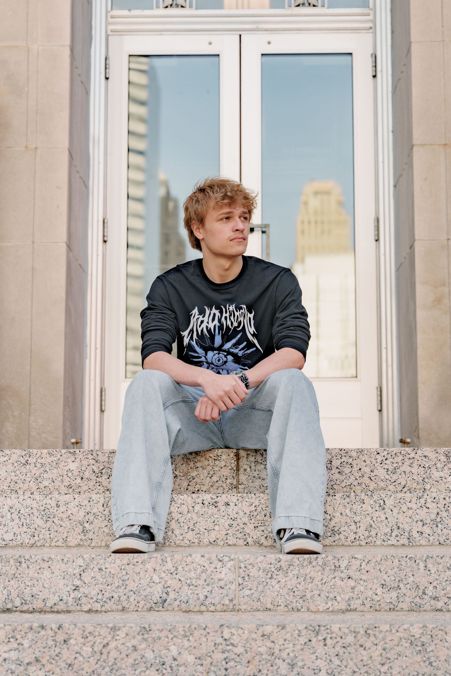 Young man in graphic sweatshirt and jeans sitting on stone steps in front of glass doors in urban setting.