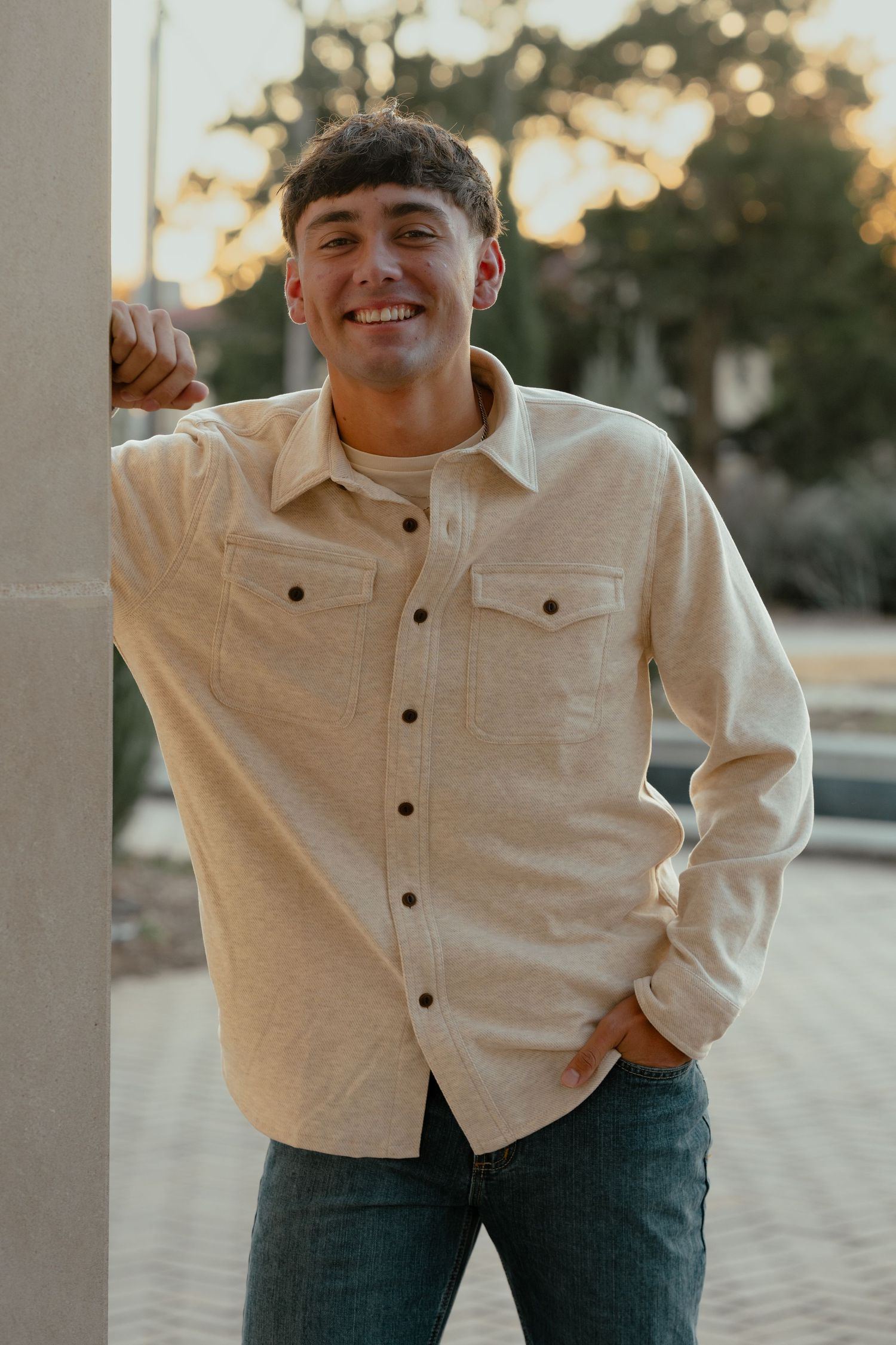 A warm-toned portrait of someone in a light button-down shirt leaning against a column at sunset.