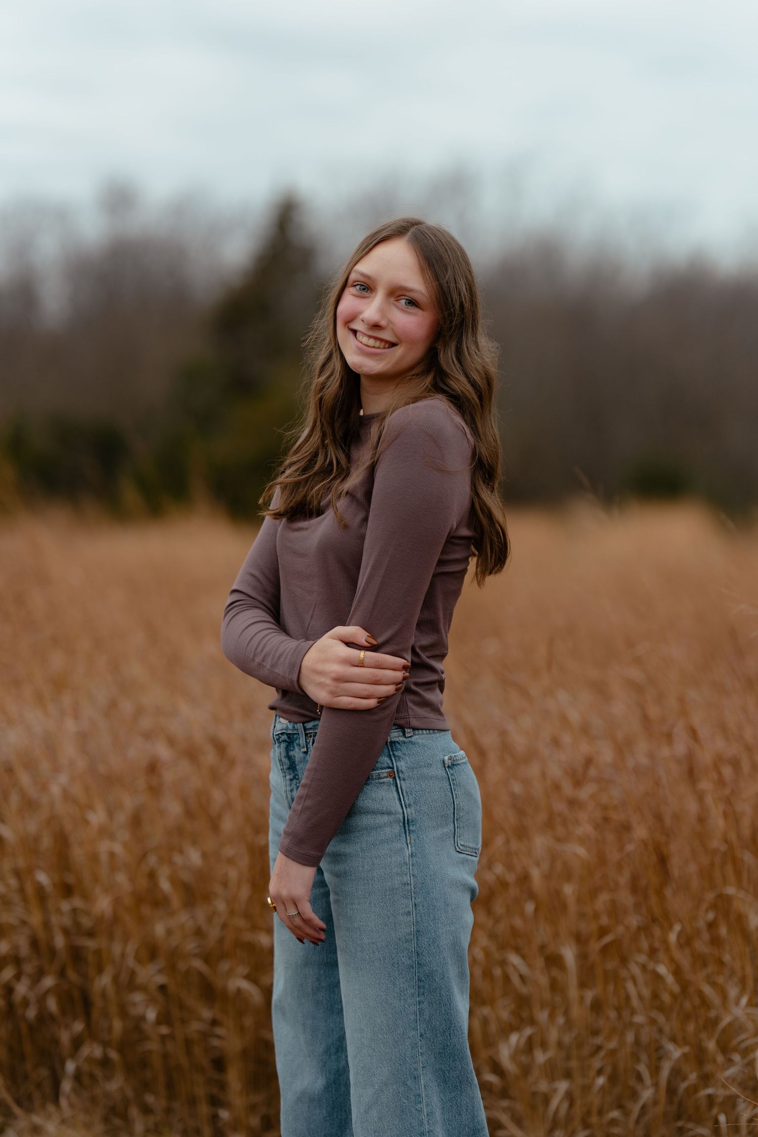 Young woman in brown top and wide-leg jeans smiling in golden field with trees in background.