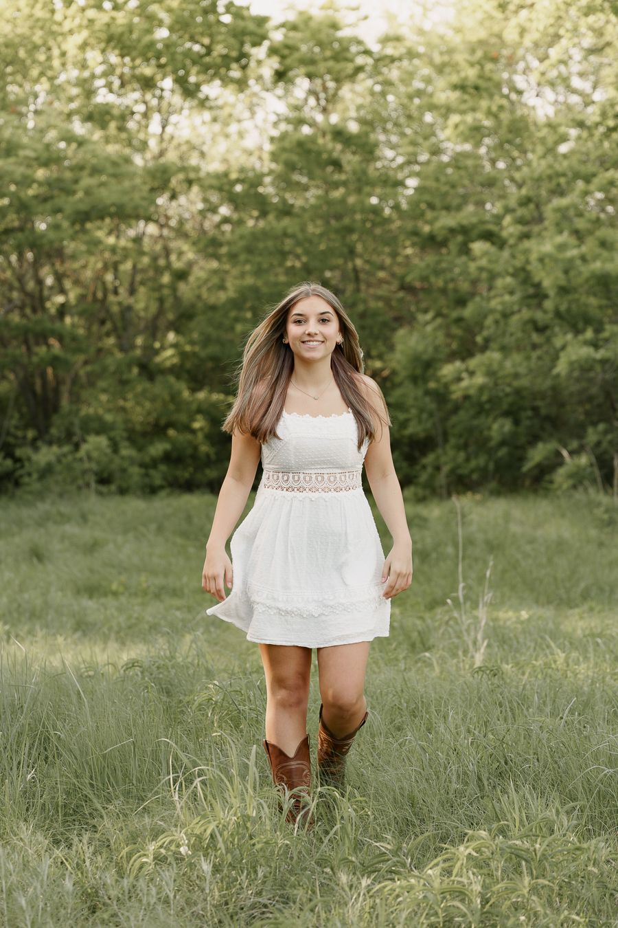 Young woman in white dress and cowboy boots walking through green meadow, smiling at camera.