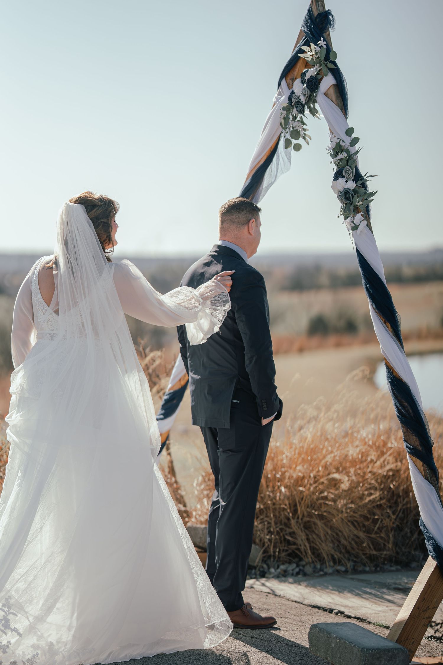 Bride and groom stand before a rustic floral arch outdoors during their wedding ceremony on a sunny day.