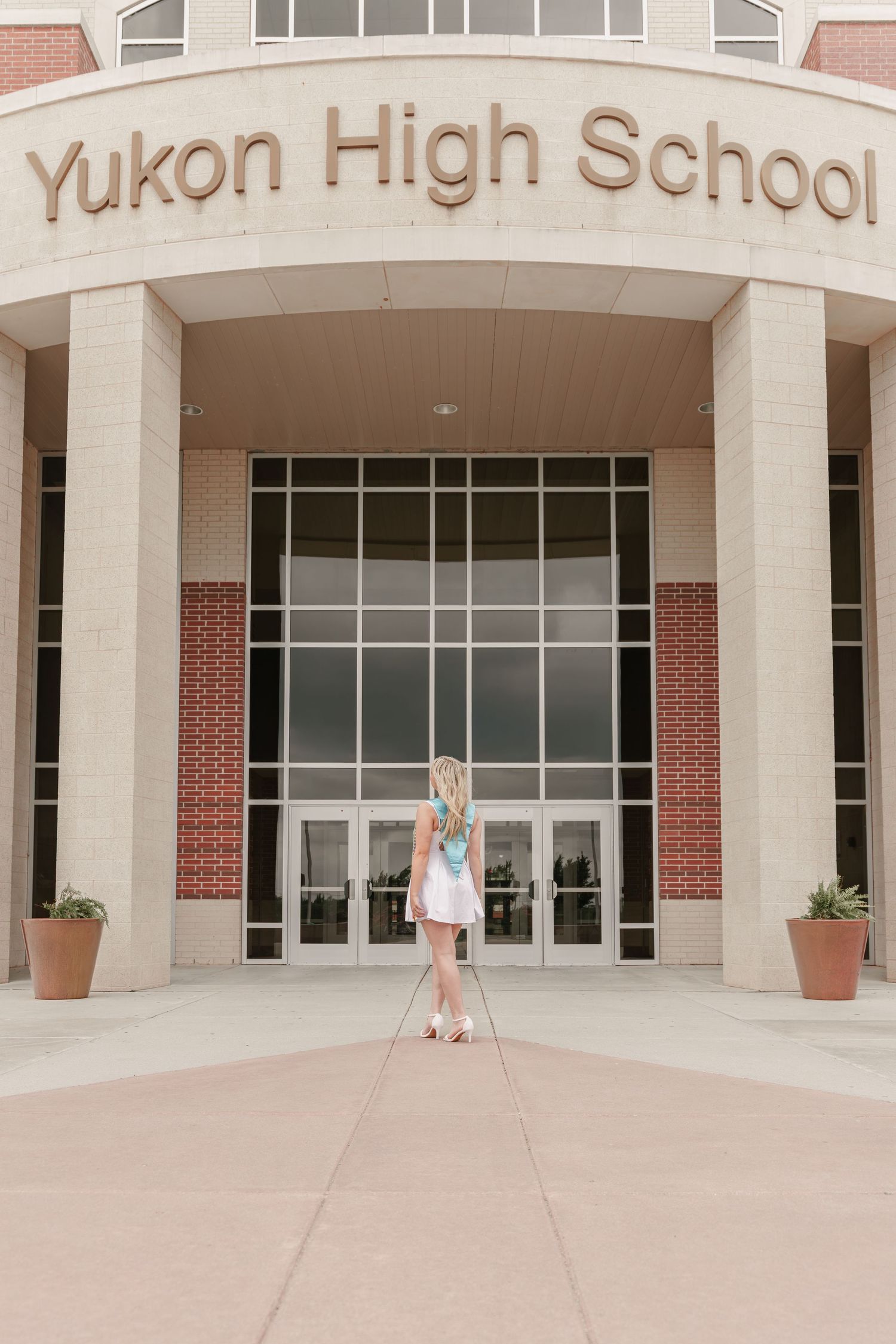 A blonde girl in a white dress stands before Yukon High School's entrance, holding a diploma.