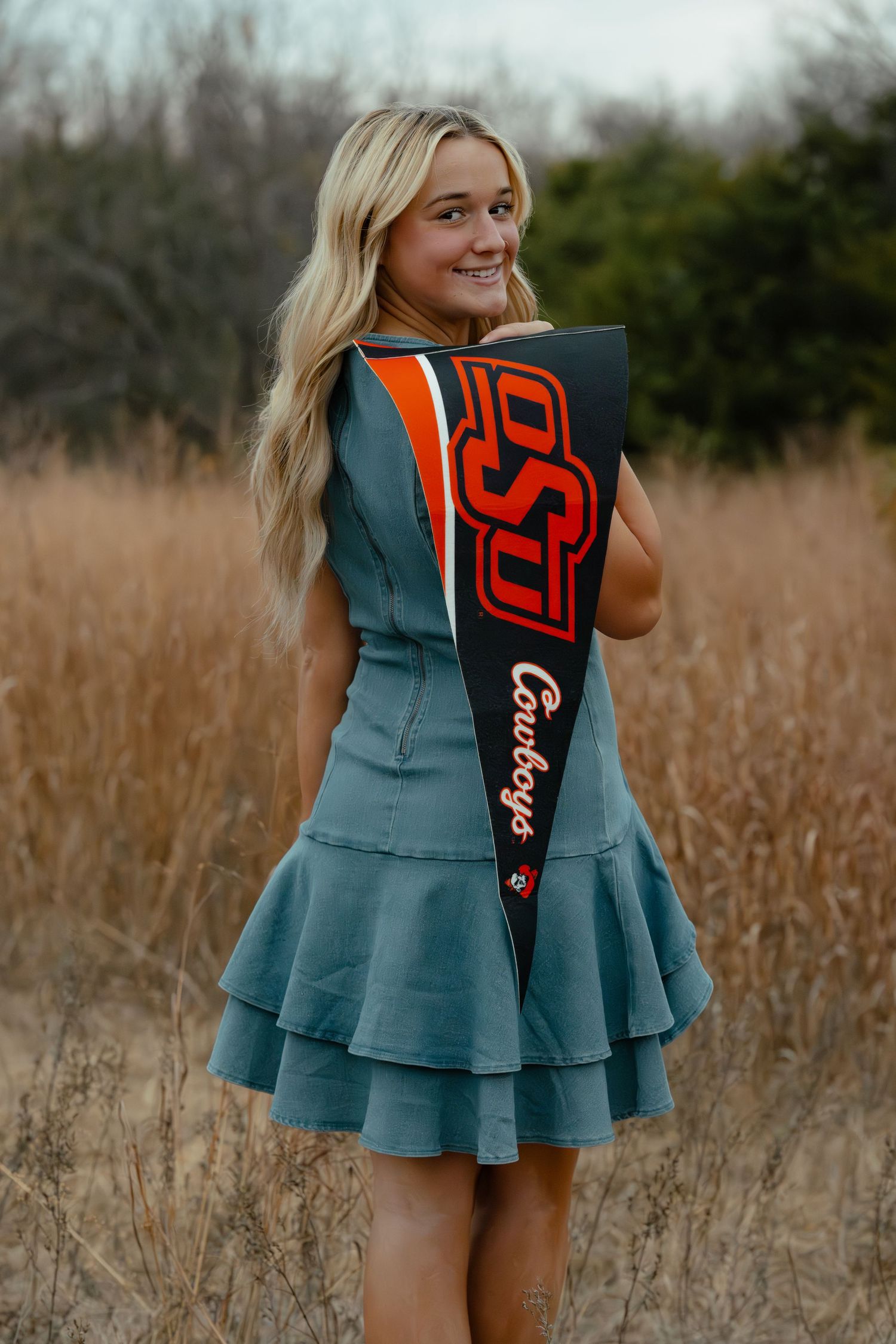 Smiling blonde woman in teal ruffled dress holding OSU Cowboys pennant flag in a field of tall dry grass.