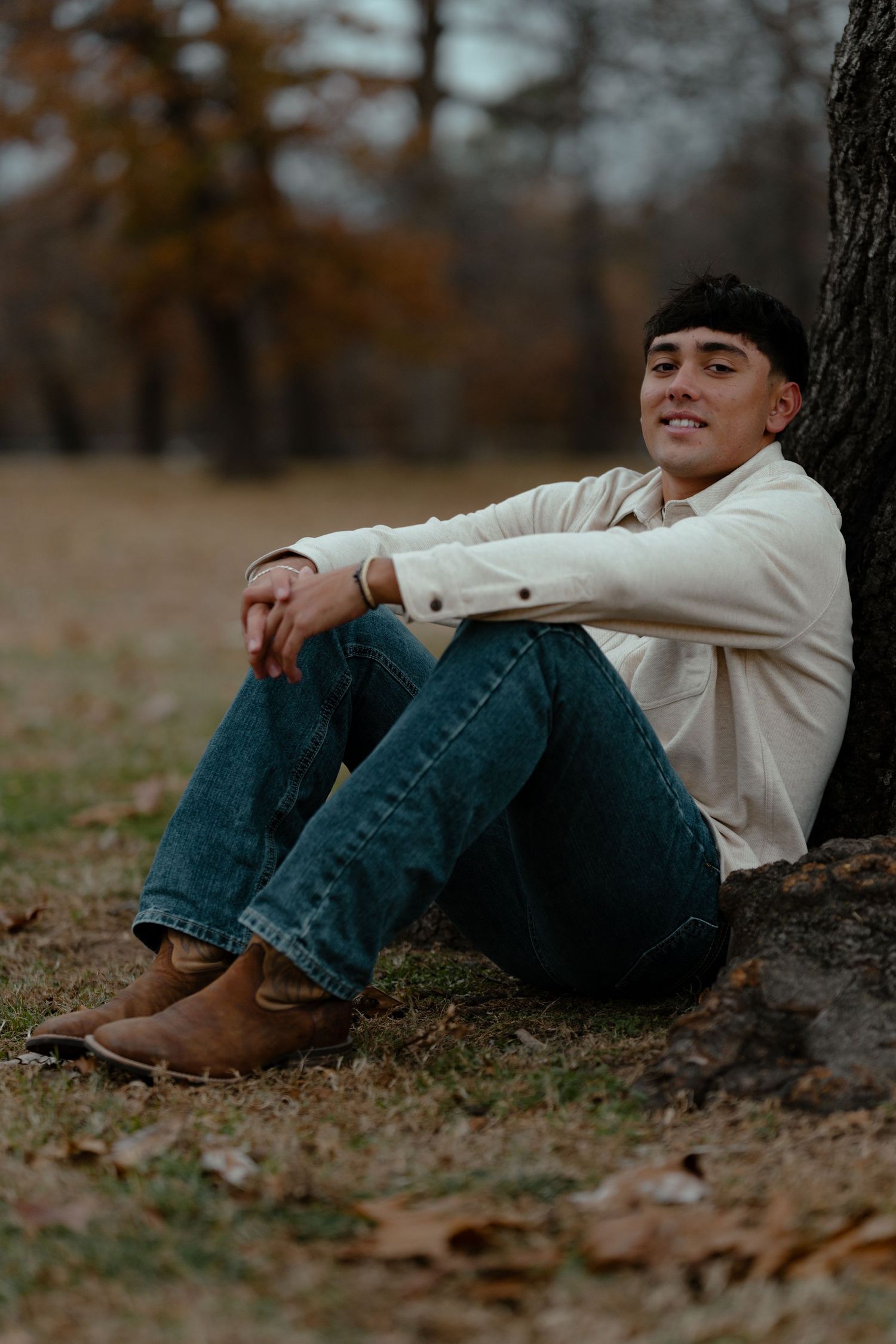 Person sitting against tree trunk wearing cream shirt and jeans during autumn season.