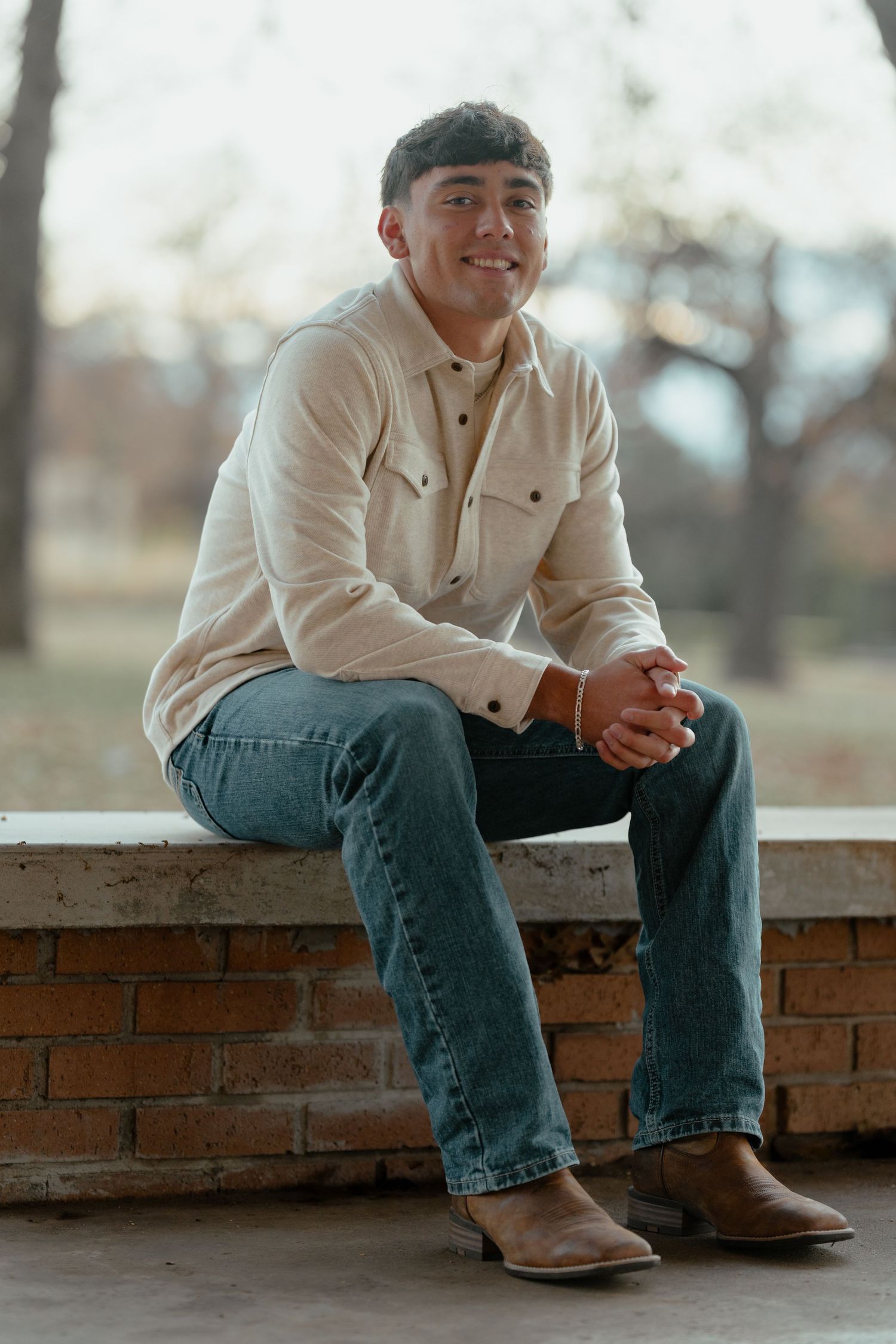 Person sitting on brick wall wearing cream button-down shirt and jeans in outdoor setting.