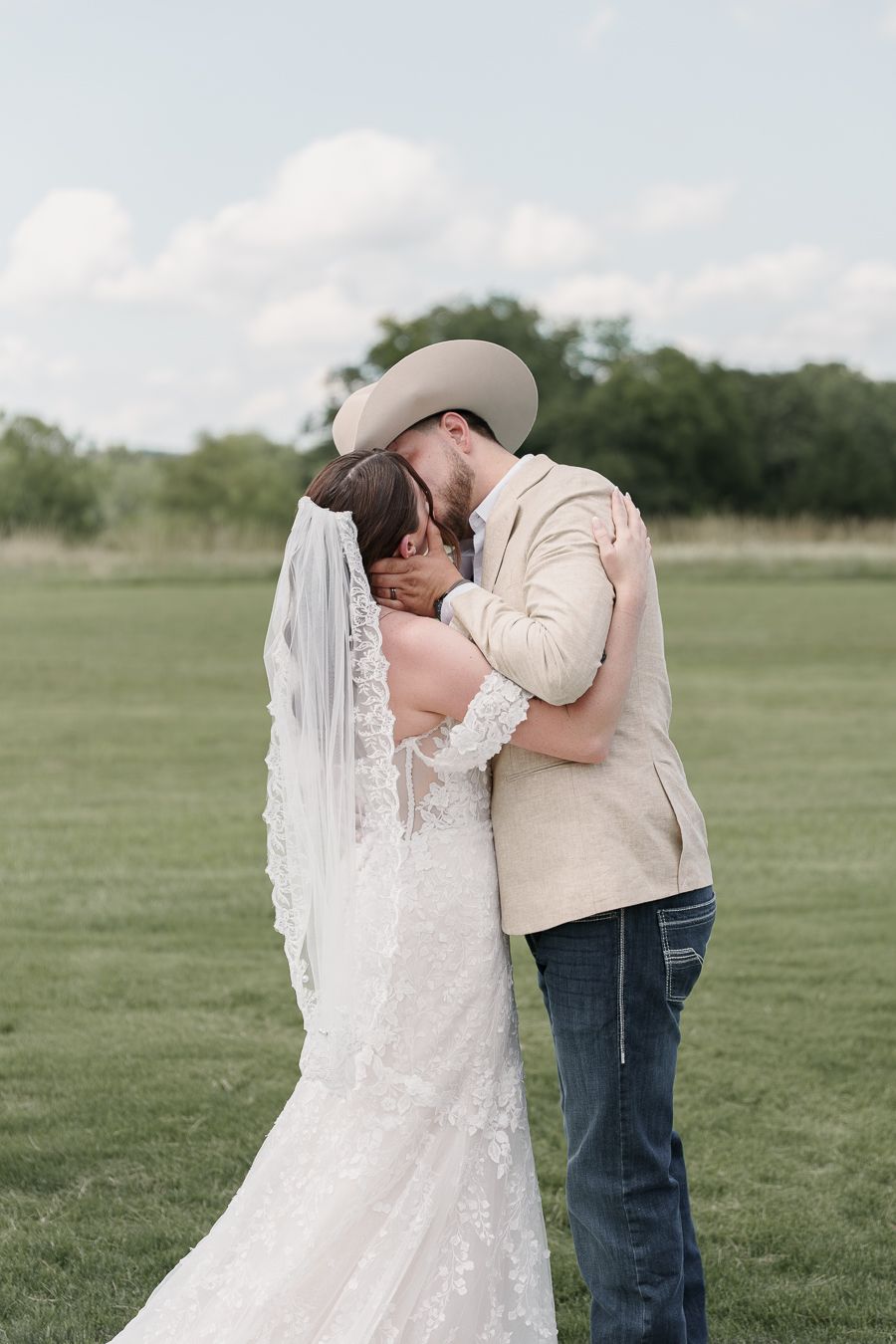 A bride and groom share a romantic kiss in a green field, the groom wearing a cowboy hat and the bride in a lace wedding gown.