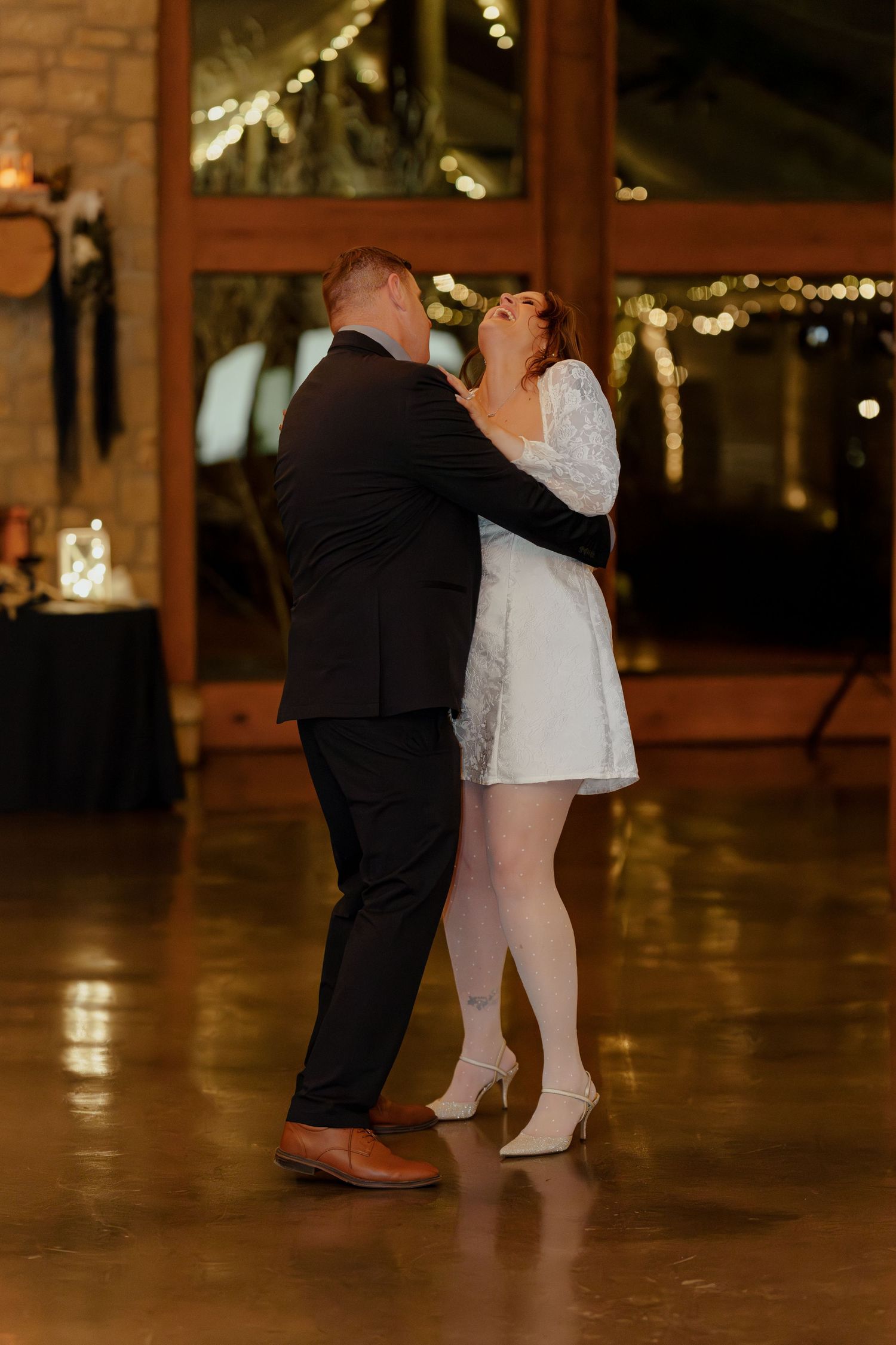 Bride in white mini dress and groom in black suit share their first dance at a warmly lit wedding reception venue.