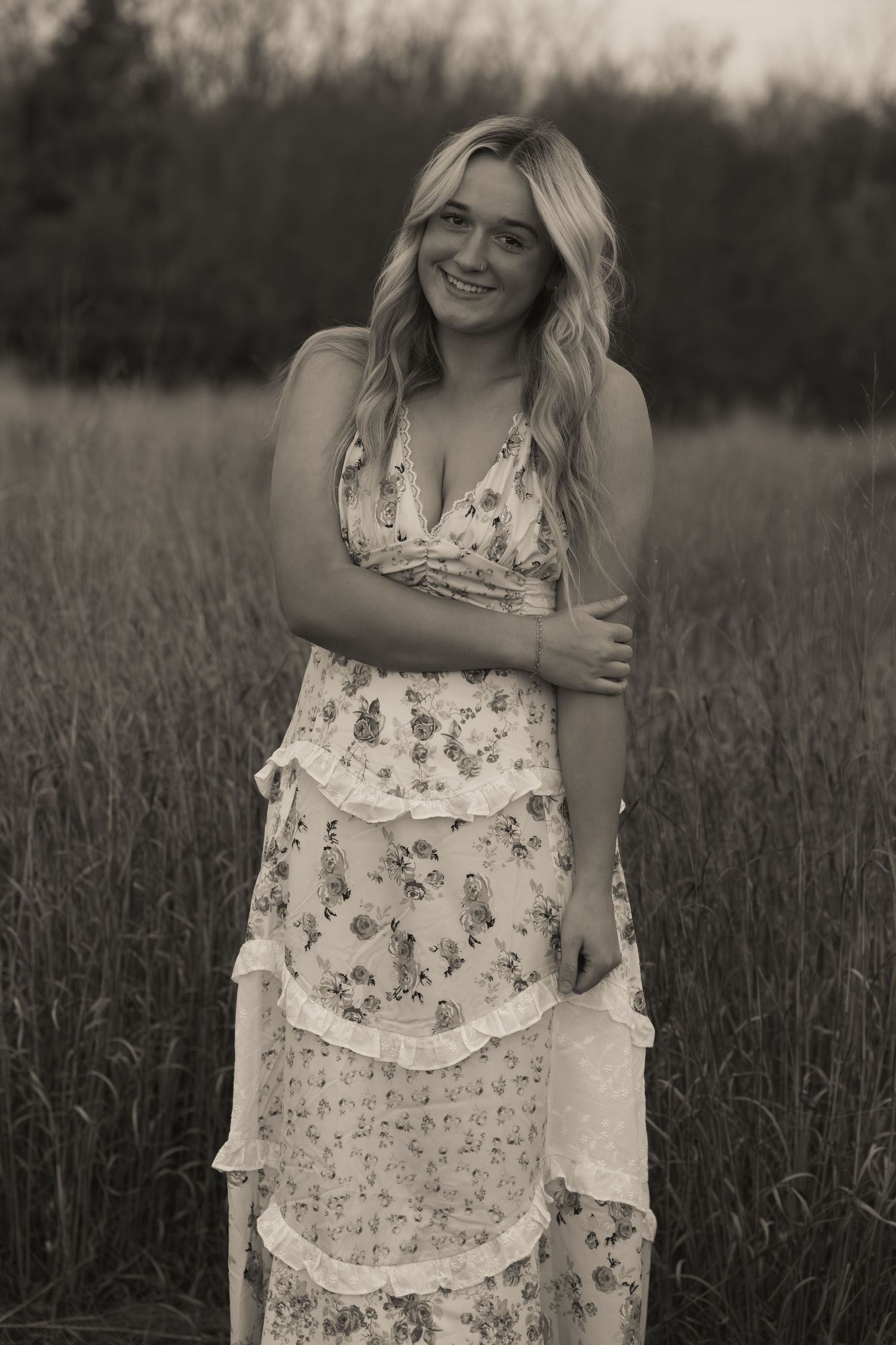 Smiling blonde woman in floral maxi dress standing in tall grass field, black and white portrait photography.
