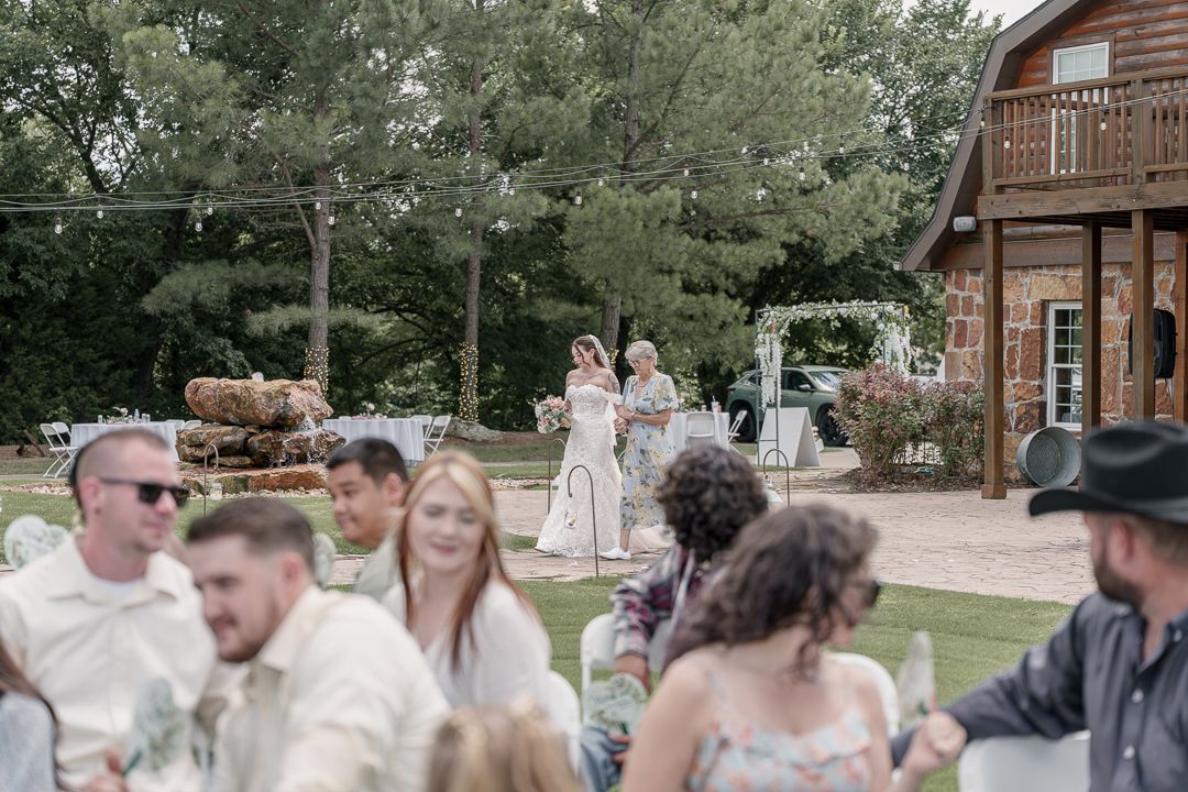 Outdoor wedding reception at rustic barn venue with guests seated on lawn, bride at altar, string lights, and lush green trees.