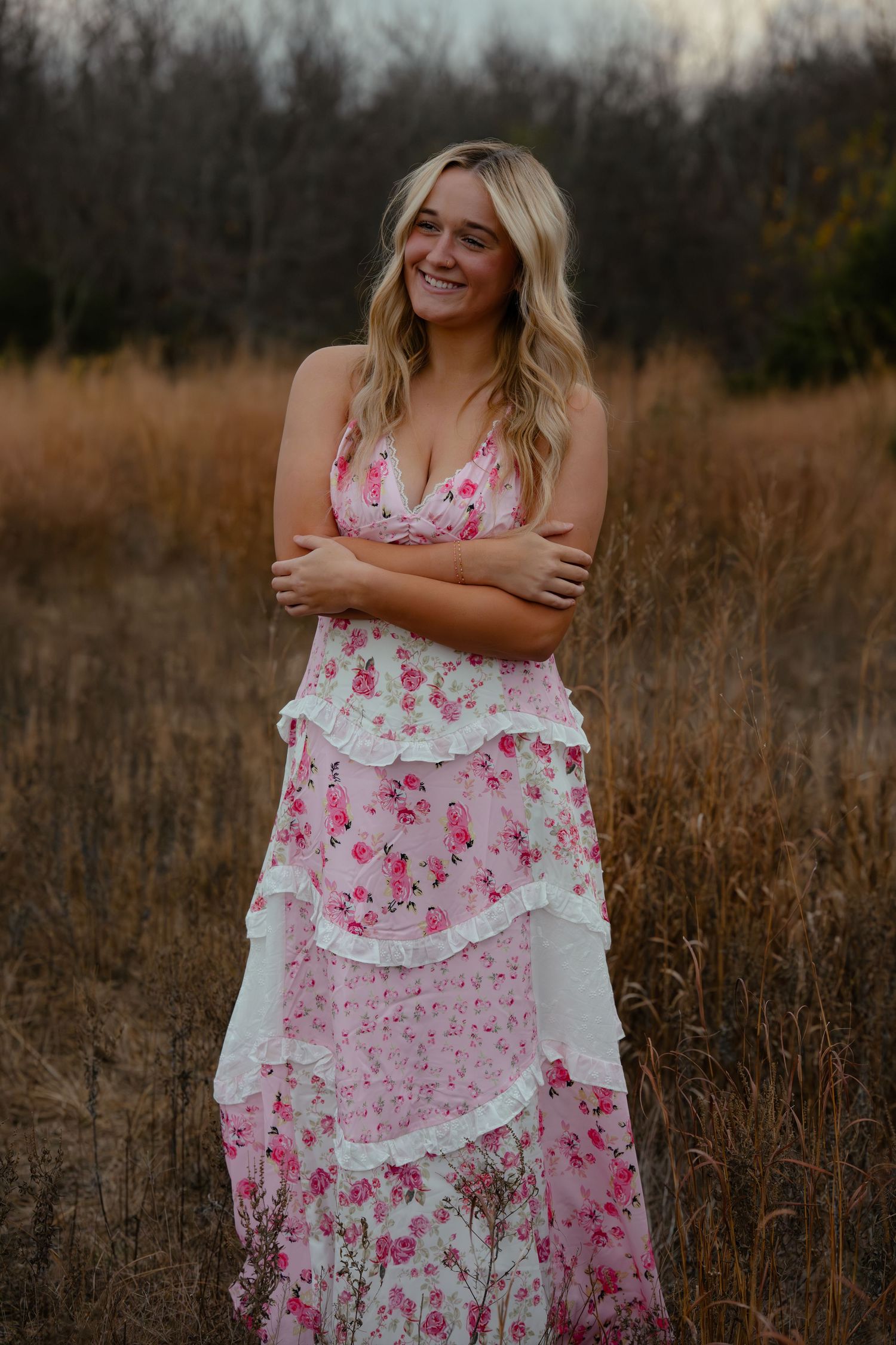 Smiling blonde woman in a pink floral tiered ruffle maxi dress standing in a golden field at dusk.