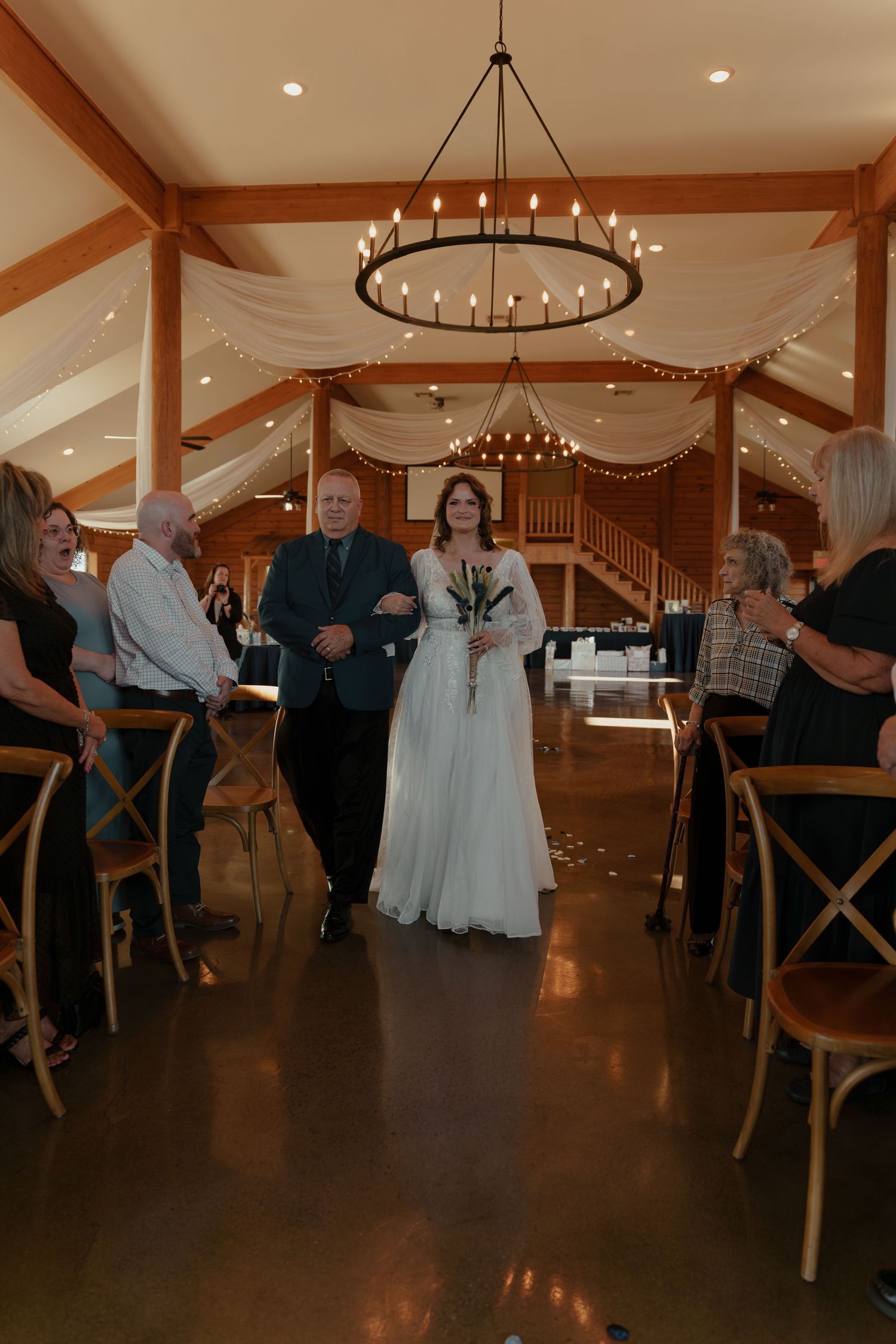 A wedding ceremony takes place in an elegant barn venue with wooden beams and circular chandeliers overhead.