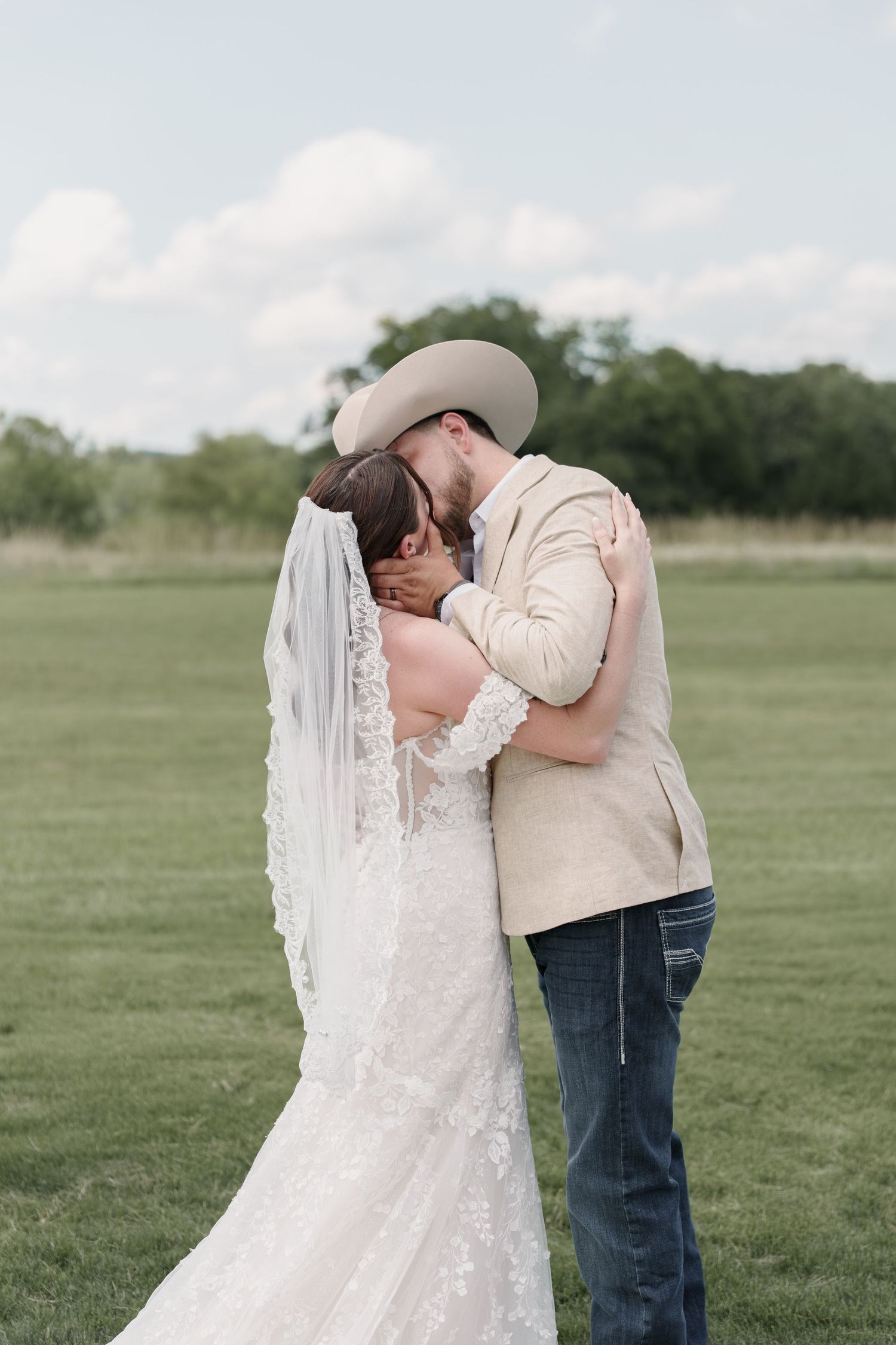 A bride and groom share a romantic kiss in a green field, with the bride in lace gown and groom in cowboy hat.