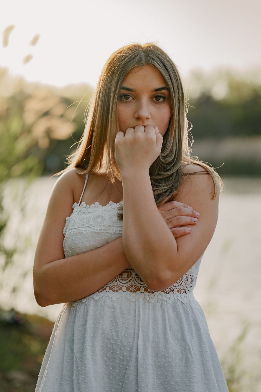 Young woman in white lace sundress standing near a lake at golden hour, looking pensively at the camera.