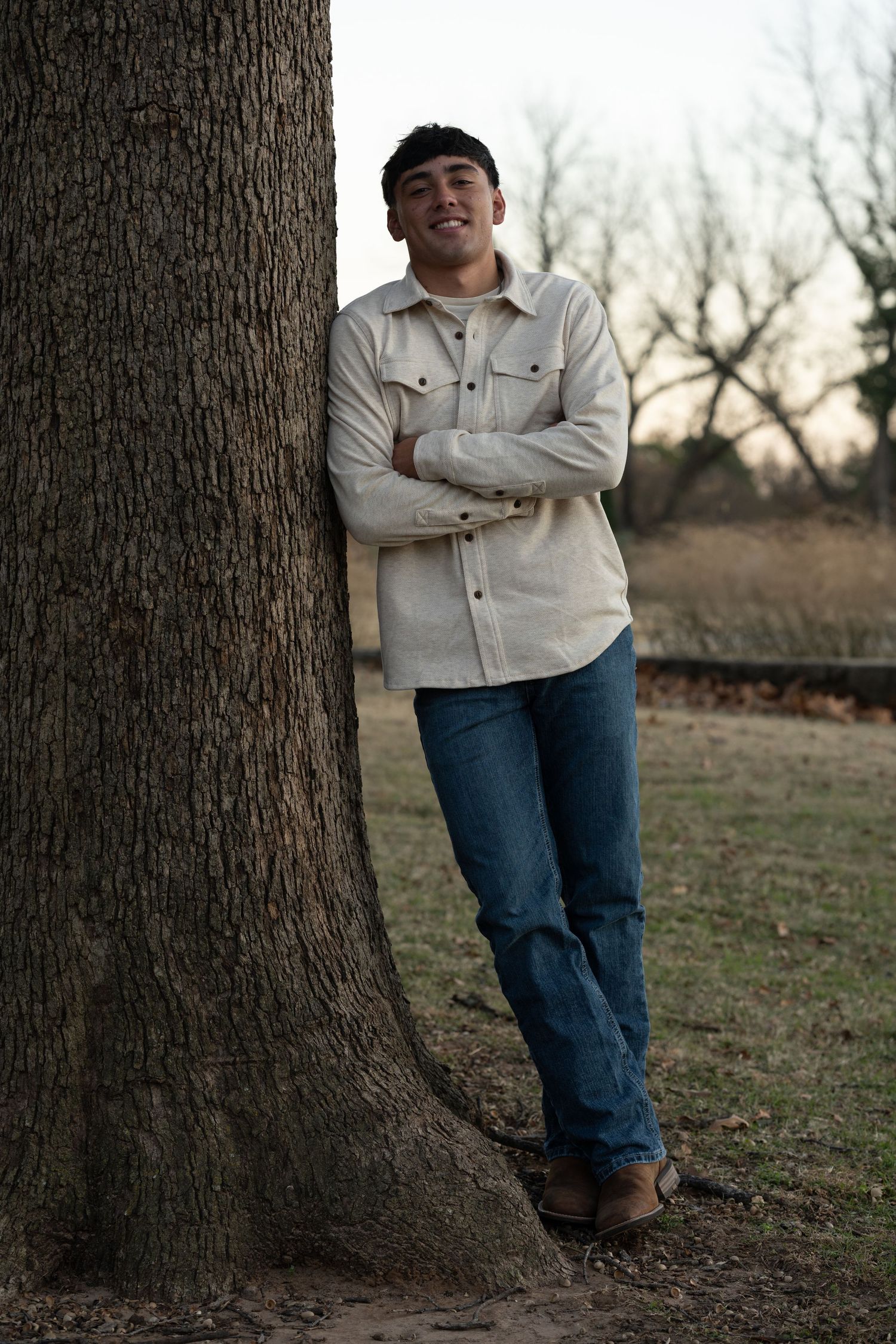 Person leaning against a large tree trunk wearing a light-colored jacket and blue jeans.