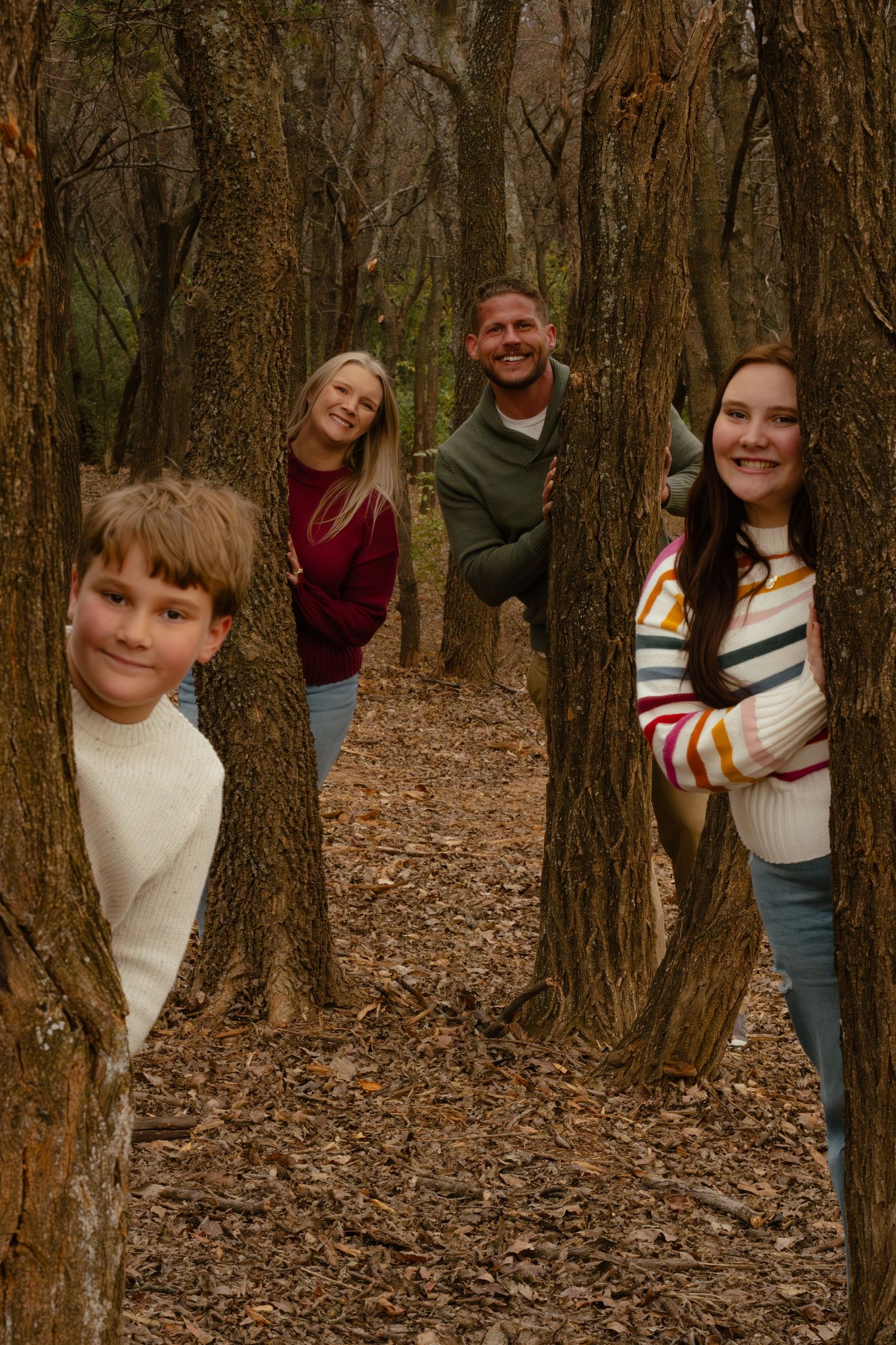 Playful family peeking from behind trees in a wooded autumn setting.