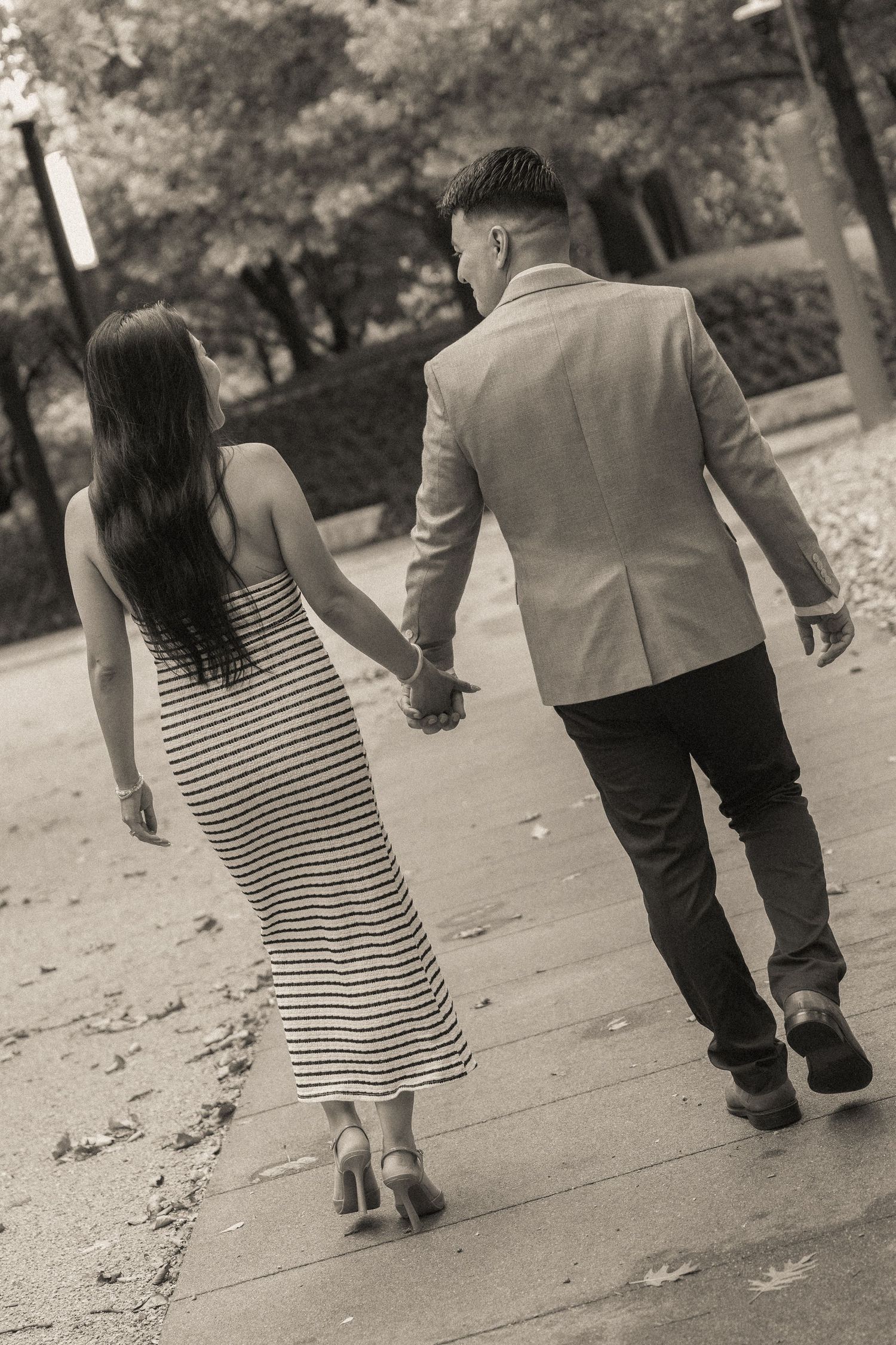 Black and white photo of a couple holding hands walking down a path in a park, woman in striped dress and heels.