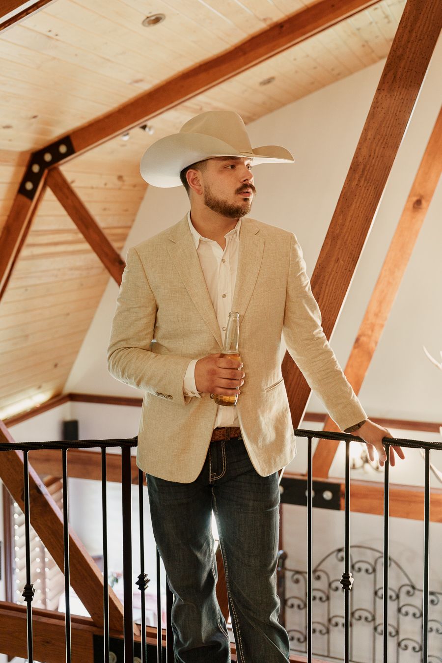 Man in cowboy hat and tan blazer holding champagne glass on rustic wooden staircase.
