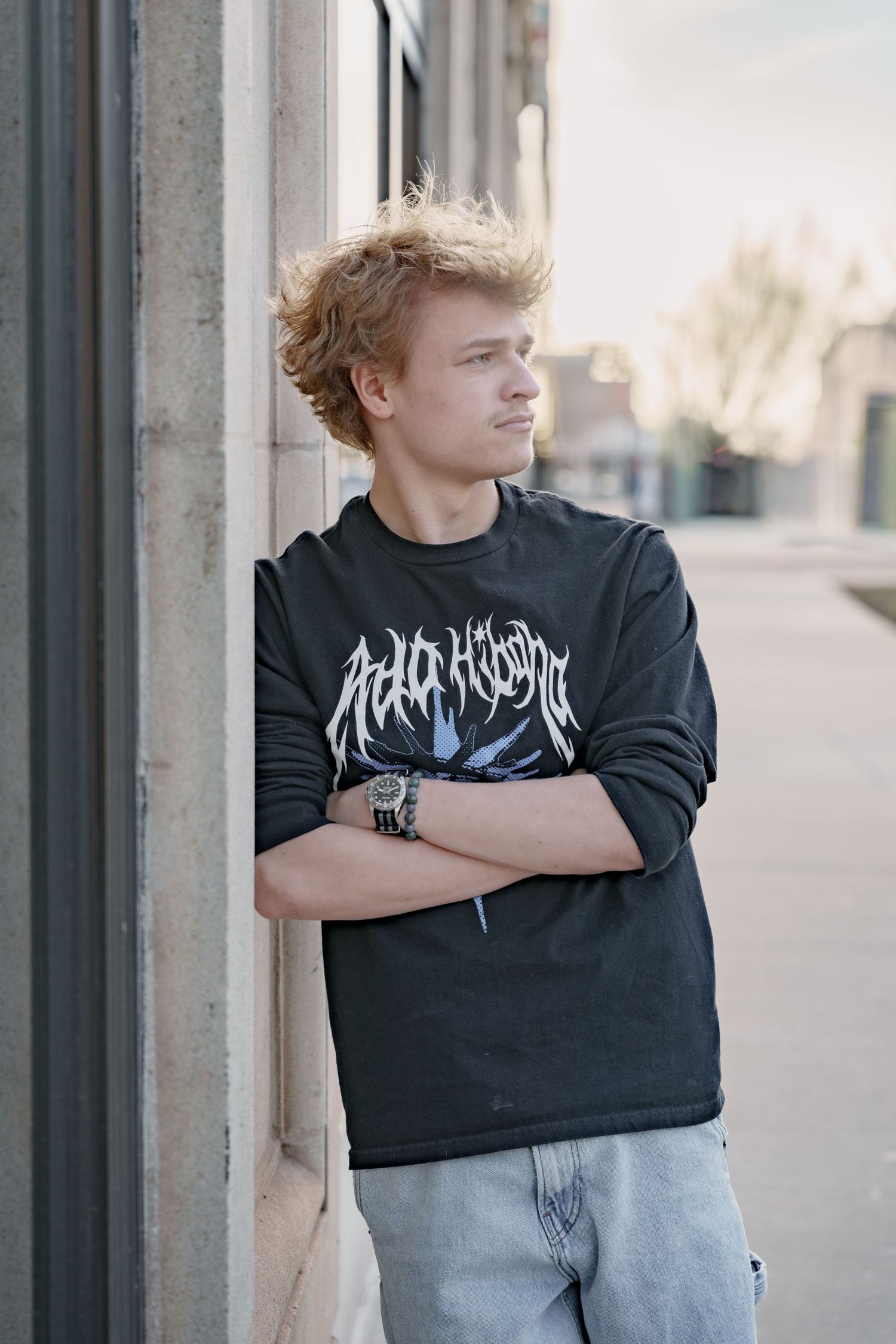 Young man with curly blonde hair in black graphic long-sleeve shirt leaning against building wall with arms crossed.